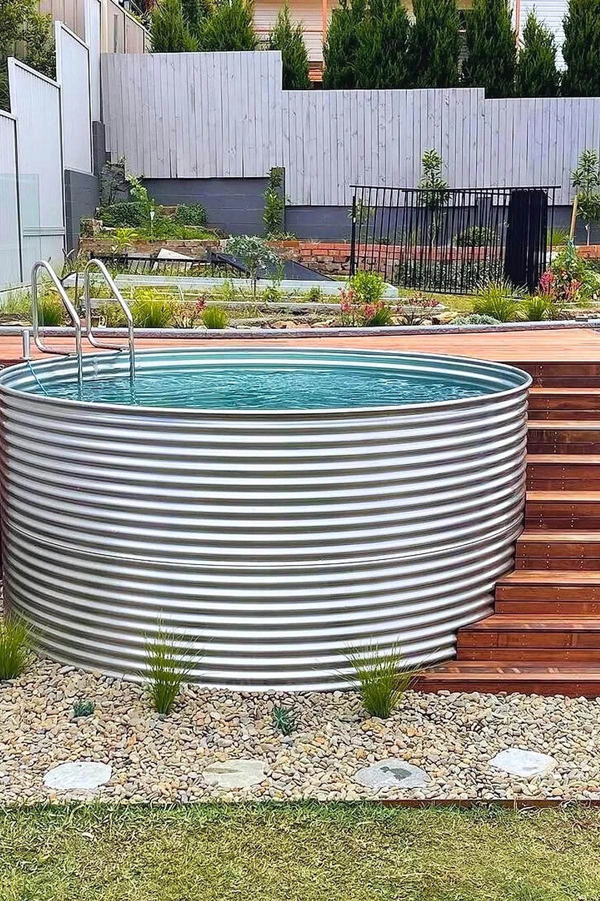 A small round pool made of silver corrugated metal with wooden steps on the side, surrounded by gravel and small plants, set in a backyard with a white fence and green lawn.