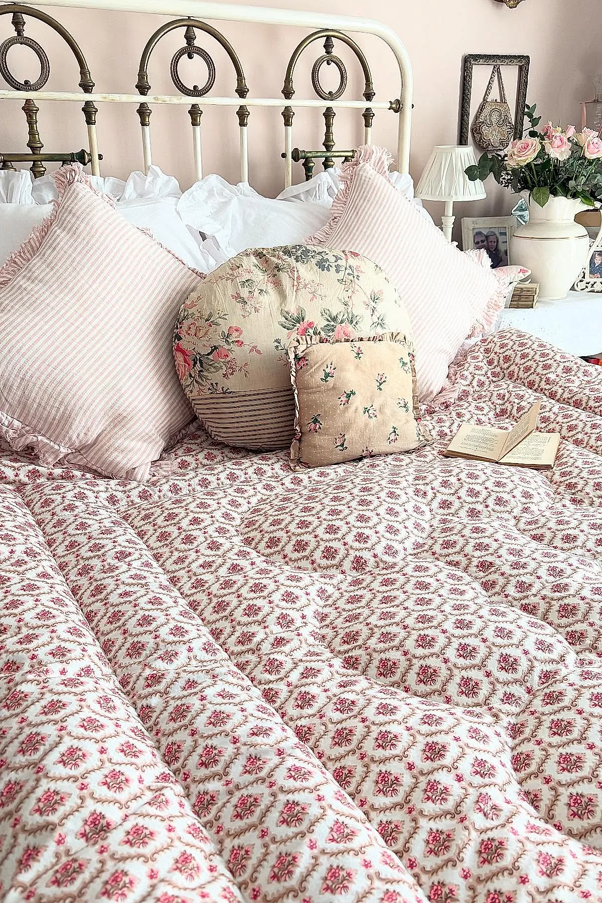 vintage bedroom featuring antique white metal bedframe with brass accents, pink striped ruffled pillows, round floral patterned cushion, small square floral cushion on red and beige patterned quilt