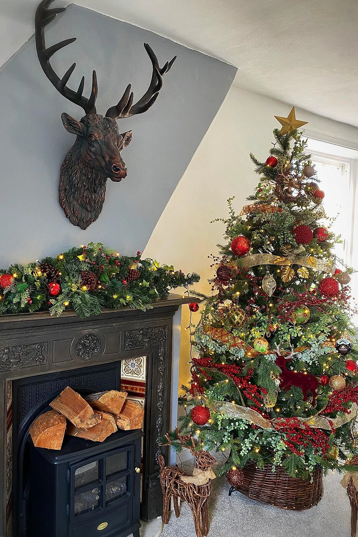 Christmas tree decorated with red and gold ornaments, berries, greenery, pinecones beside dark fireplace mantel adorned with matching garland and lit string lights, wicker deer sculptures on carpeted floor under mounted deer head