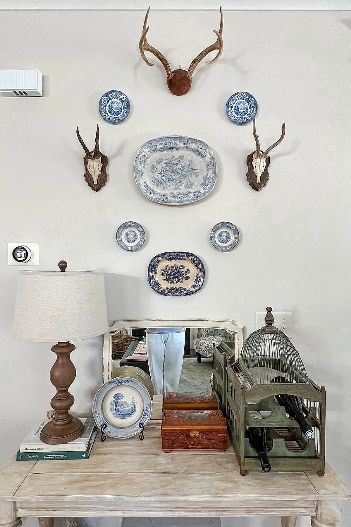 Living room wall decor featuring mounted antlers and skulls alongside various blue and white decorative plates arranged on a light gray wall above a distressed wooden table with a brown lamp, vintage wooden boxes, and an antique wine rack.
