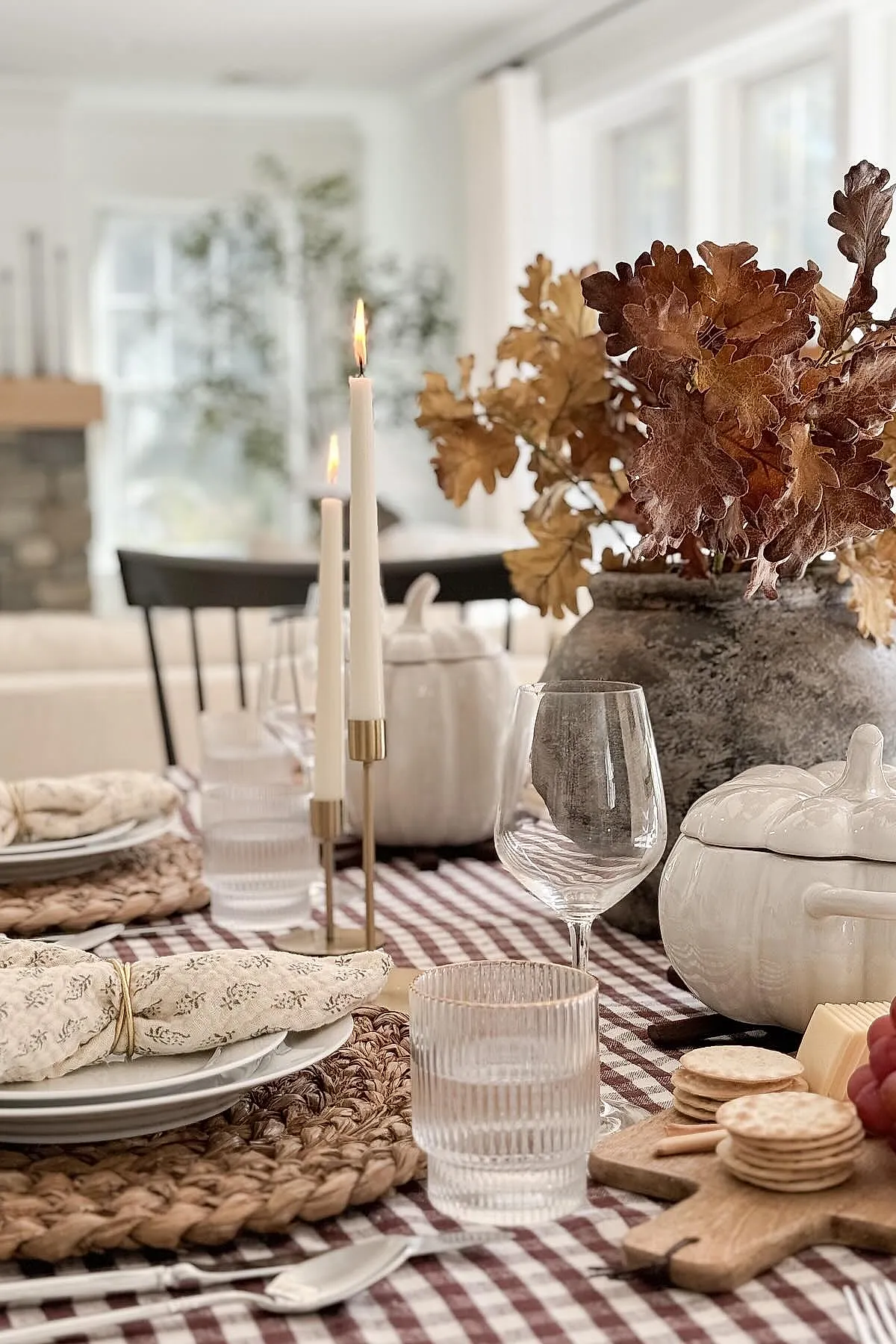 Thanksgiving table setting with brown and white checkered tablecloth, woven round placemats, white plates with patterned napkins, two lit white taper candles in gold holders, ceramic white pumpkin containers, wine glass, water glass, wooden cutting board with crackers and grapes