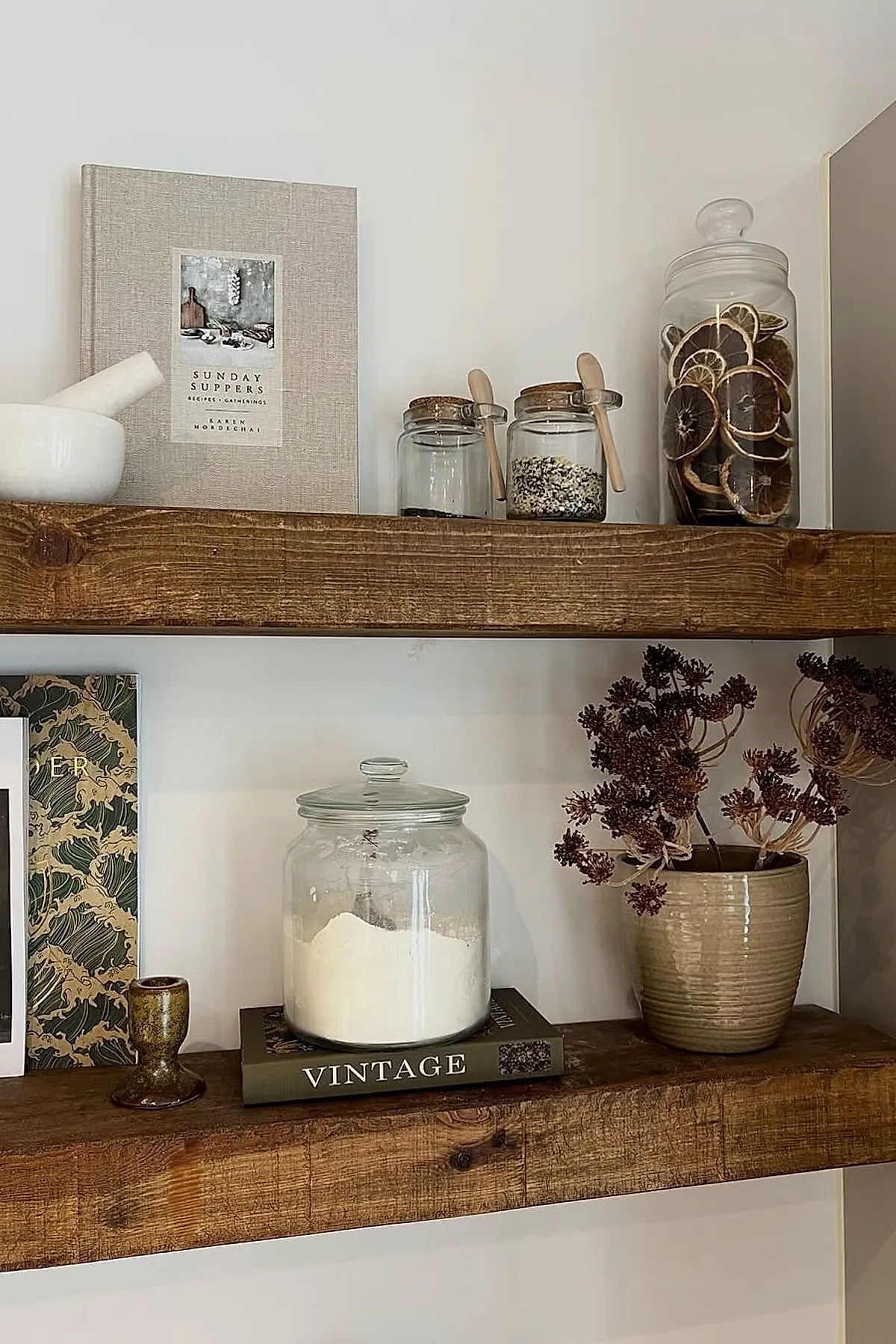 Rustic kitchen open shelves made of thick wood holding glass jars with dried citrus slices and grains, a white mortar and pestle, a beige hardcover cookbook titled