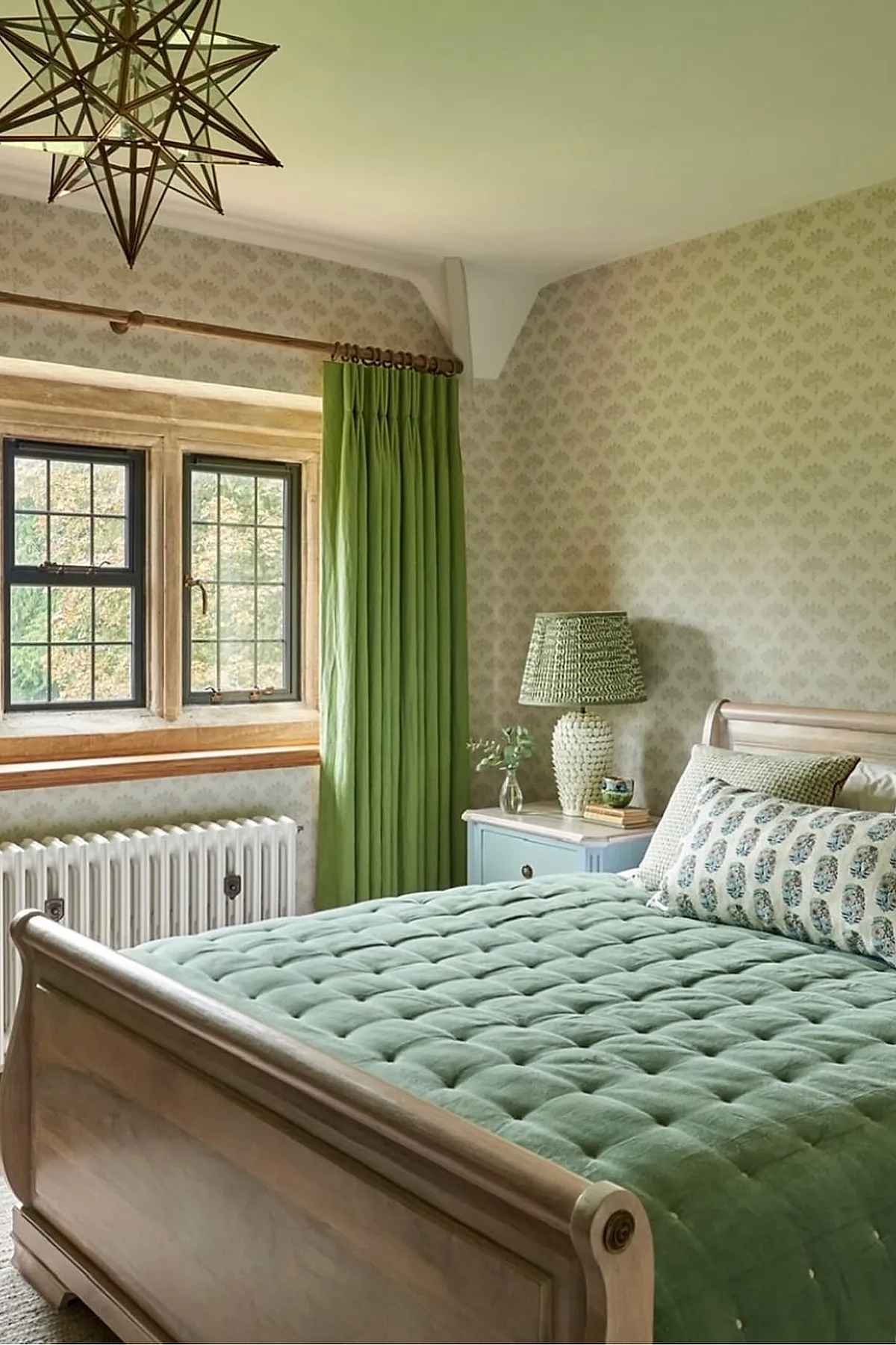 English cottage bedroom featuring vintage wooden sleigh bed with tufted sage green bedding, green floor-length curtains, patterned beige wallpaper, blue nightstand with textured lamp, and geometric star-shaped pendant light.