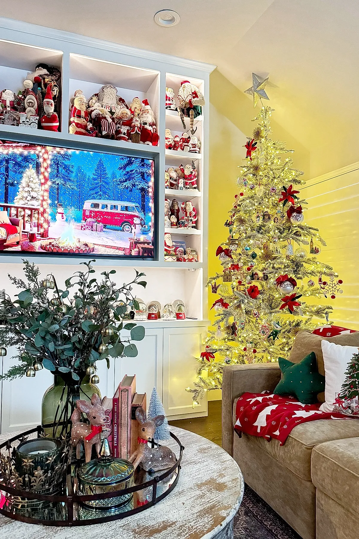christmas living room featuring a bright lit christmas tree decorated with red bows and ornaments next to a beige sofa with red and green pillows and throw blanket; white built-in shelves filled with numerous santa figurines, tv screen showing snowy winter scene, rustic round coffee table with greenery and decorative reindeer figurines