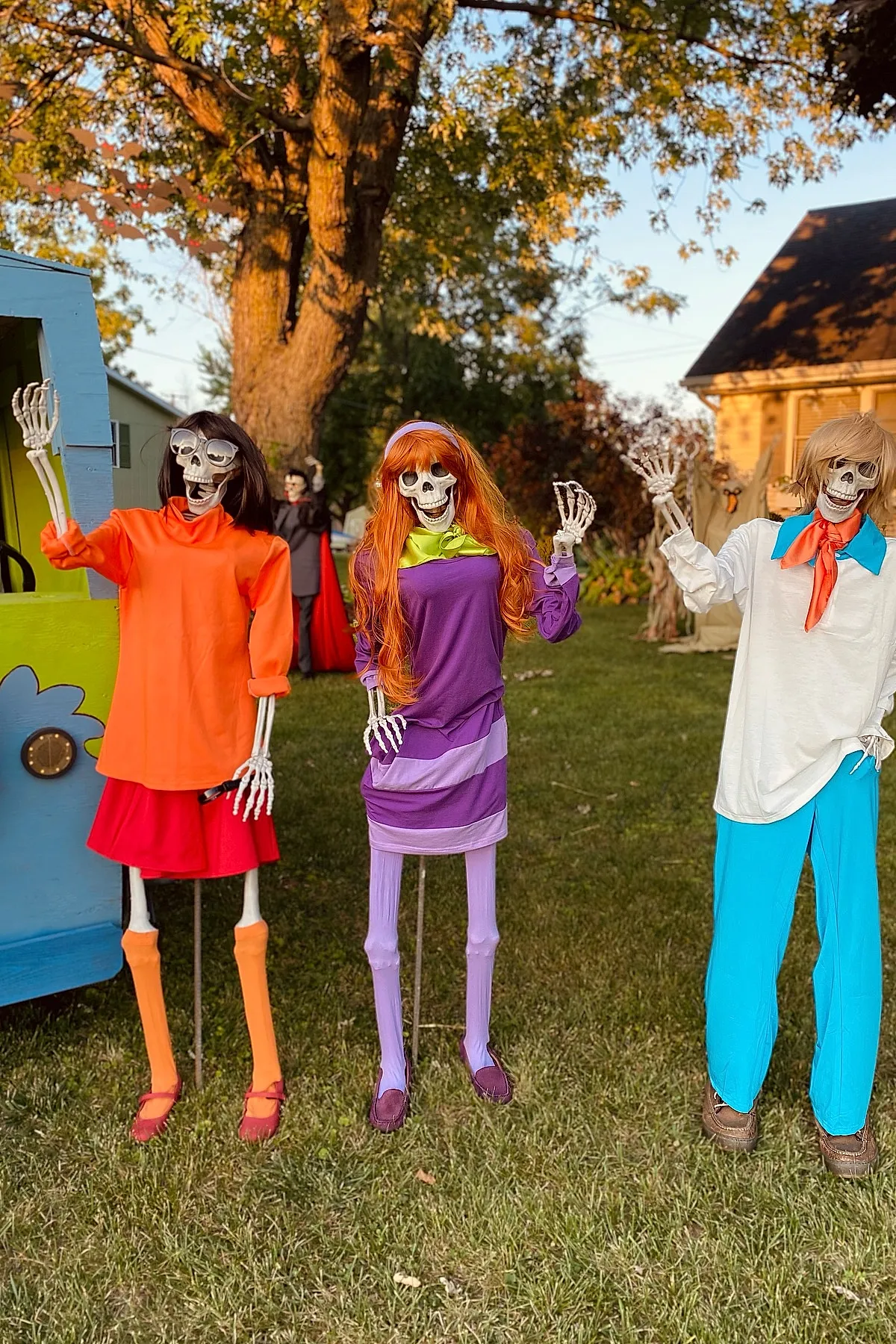Three skeleton figures outdoors wearing costumes resembling Velma in orange sweater and red skirt, Daphne in purple dress with red hair, and Fred in white shirt with blue pants and orange ascot standing on green grass near a painted blue and green van.