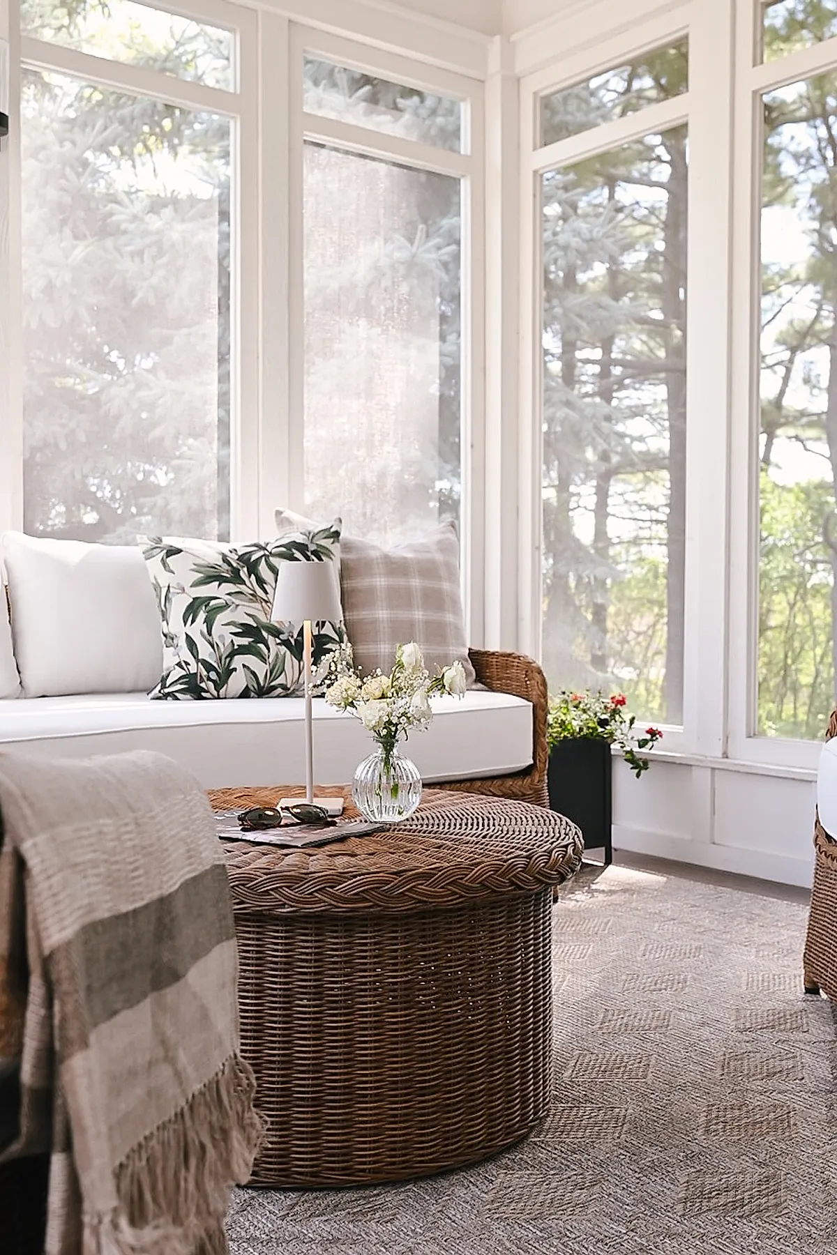 Cozy screened in porch featuring wicker sofa with white cushions and patterned pillows, round wicker coffee table with glass vase of flowers, large screened windows showing trees outside