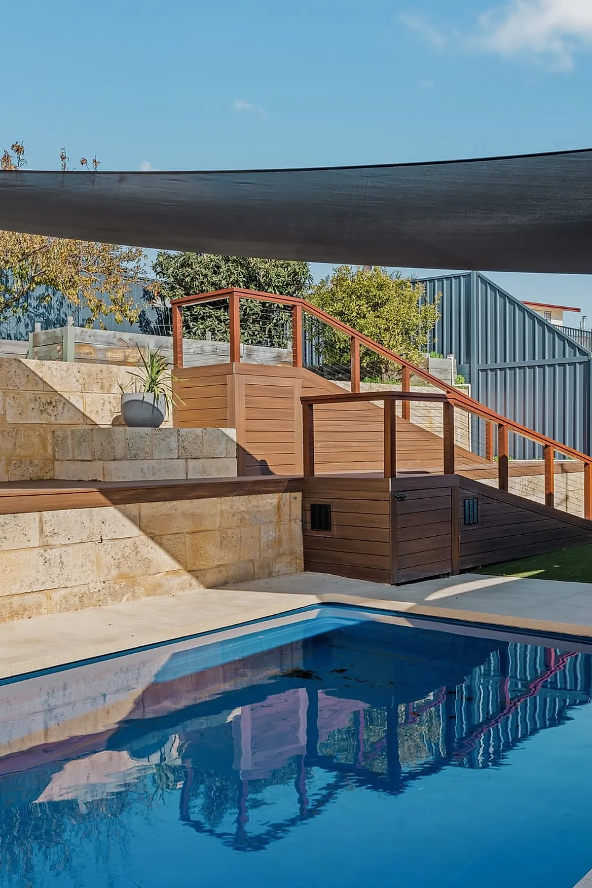 A backyard pool with a concrete deck, surrounded by stone retaining walls and wooden railings, shaded by a large black shade sail under a clear blue sky.