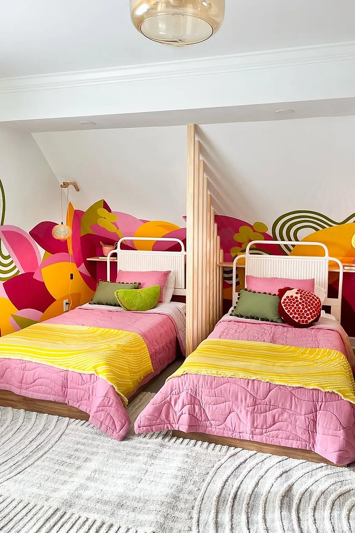 Girls shared bedroom with two white metal beds dressed in pink quilts and yellow blankets, separated by a wooden slatted divider, featuring a vibrant floral mural wall in shades of pink, orange, yellow, and green.