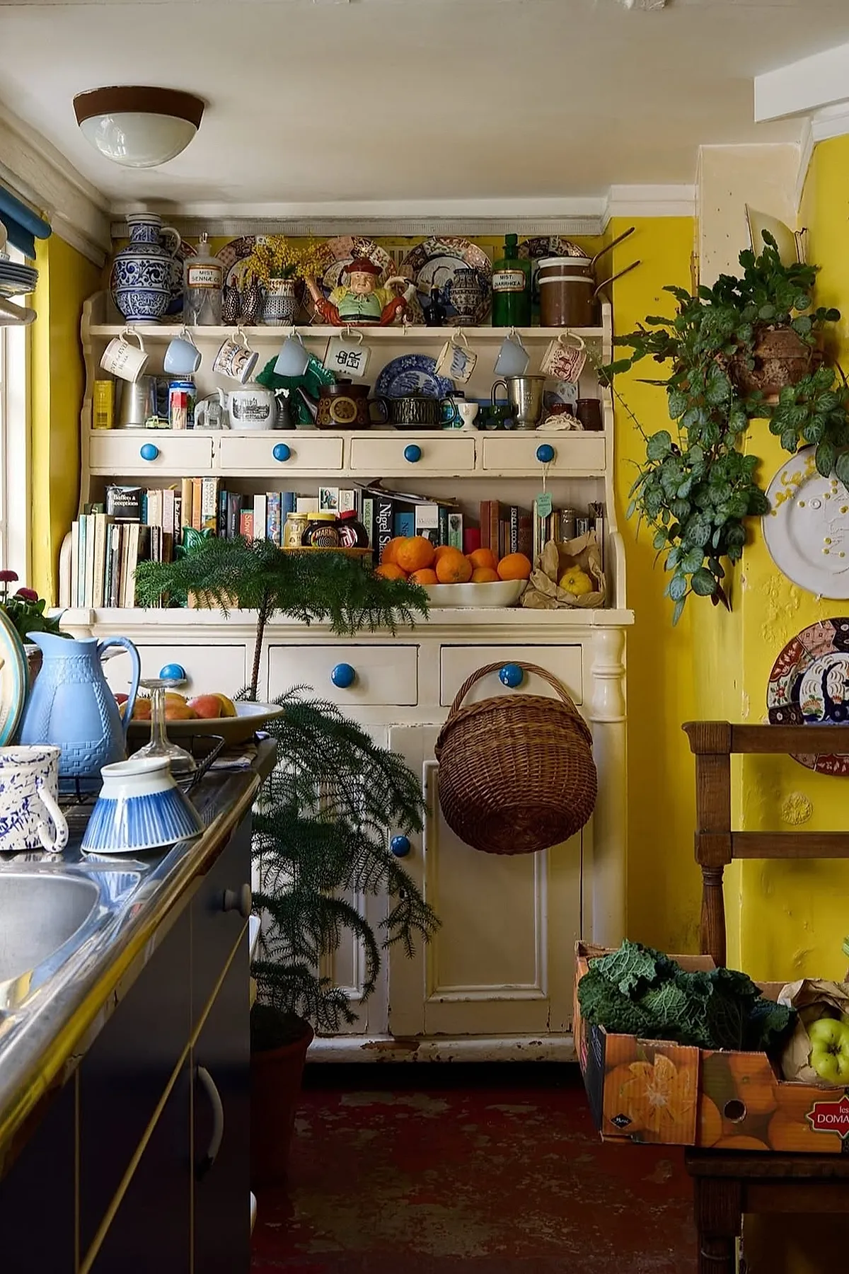 Yellow cottage kitchen with light yellow kitchen walls, a vintage white wooden hutch featuring blue drawer knobs, hanging mugs in pastel blues and whites, shelves filled with cookbooks and colorful pottery, a wicker basket hanging from the cabinet, and leafy green plants adding freshness to the cozy space.