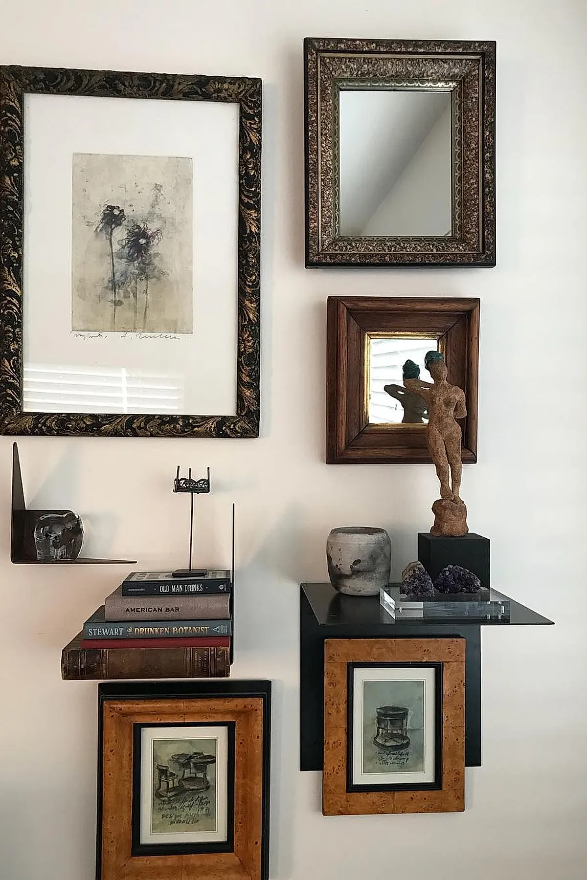 Eclectic wall decor ideas featuring vintage framed artwork and mirrors in ornate and wooden frames arranged asymmetrically on a white wall above black floating shelves holding stacked books, a small sculpture of a nude figure, amethyst geodes, and a gray ceramic pot.