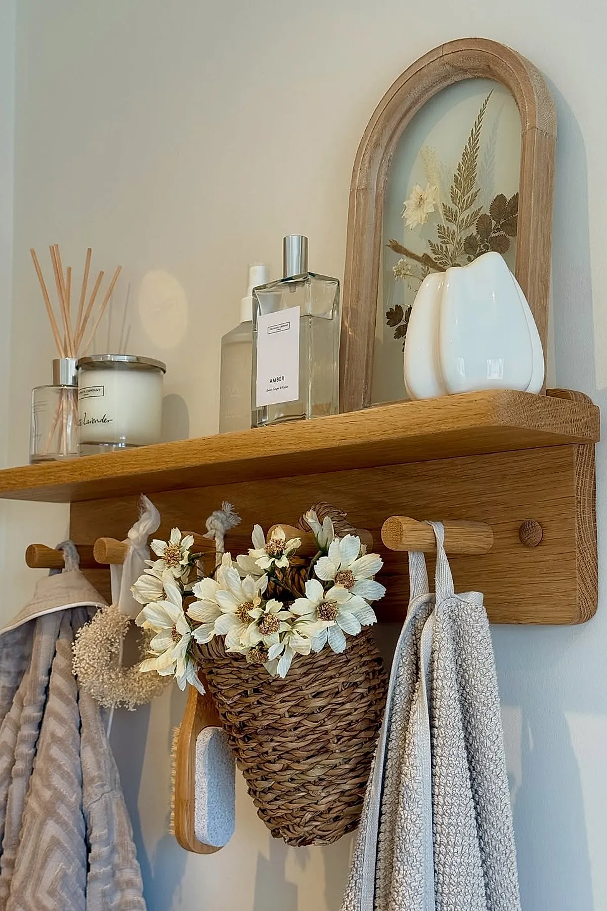 Warm neutral bathroom wooden wall shelf with pegs holding textured beige towels, a woven basket with white dried flowers, a bath brush, reed diffuser, scented candle, two bottles of perfume, and an arched wooden frame with pressed flowers against a light cream wall.