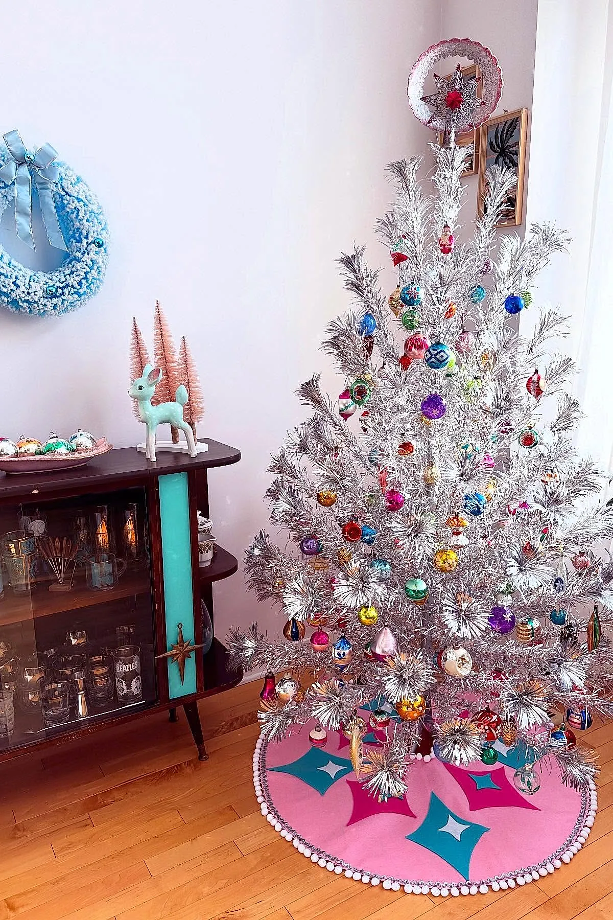 Silver vintage Christmas tree adorned with colorful glass ornaments in various shapes and patterns, set on a wooden floor with a bright pink and teal mid-century modern tree skirt beside a dark wood cabinet with turquoise accents and vintage decor.