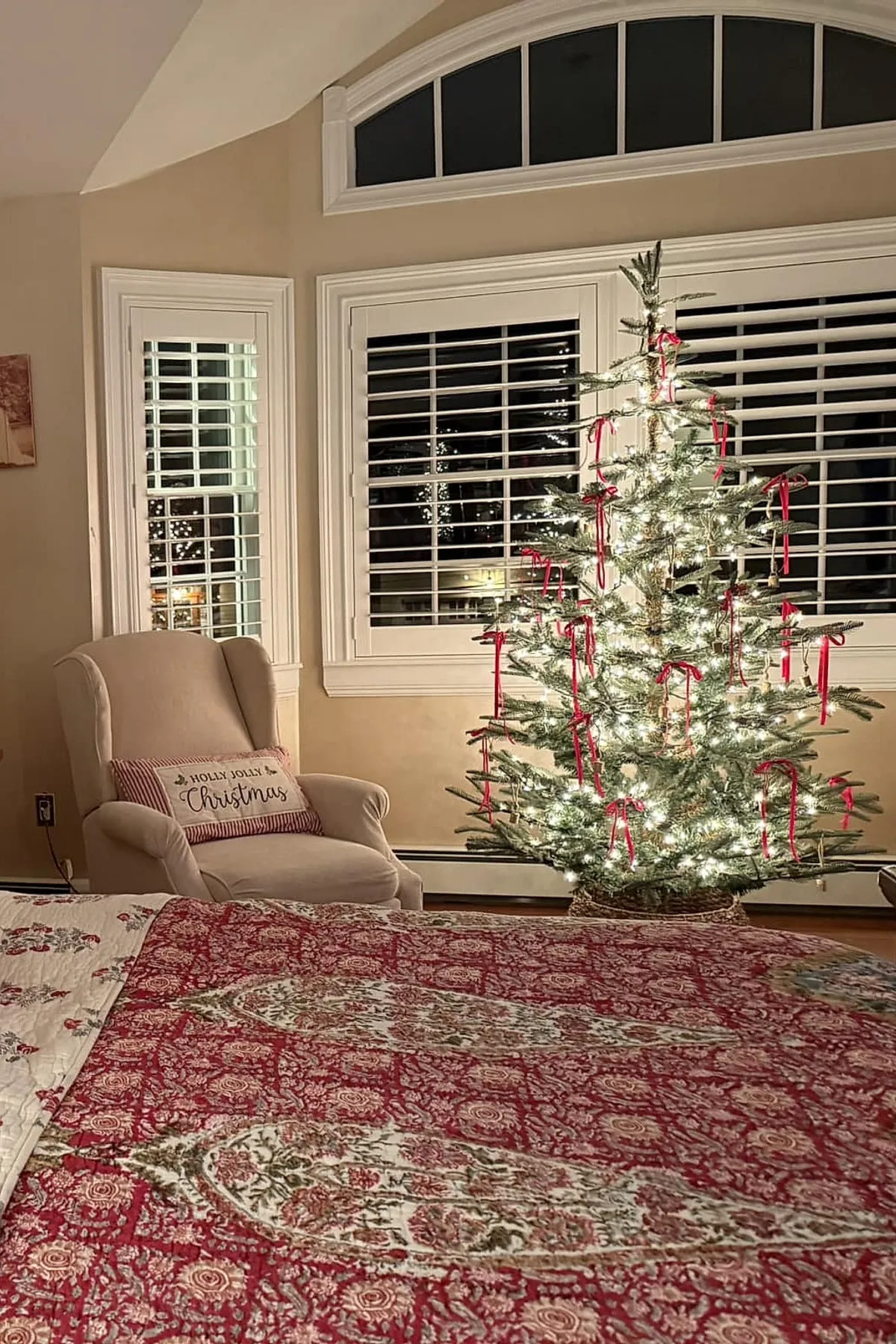 Christmas bedroom featuring a lit minimalist pine tree decorated with red ribbons, positioned near large windows with white shutters, a beige upholstered armchair with a