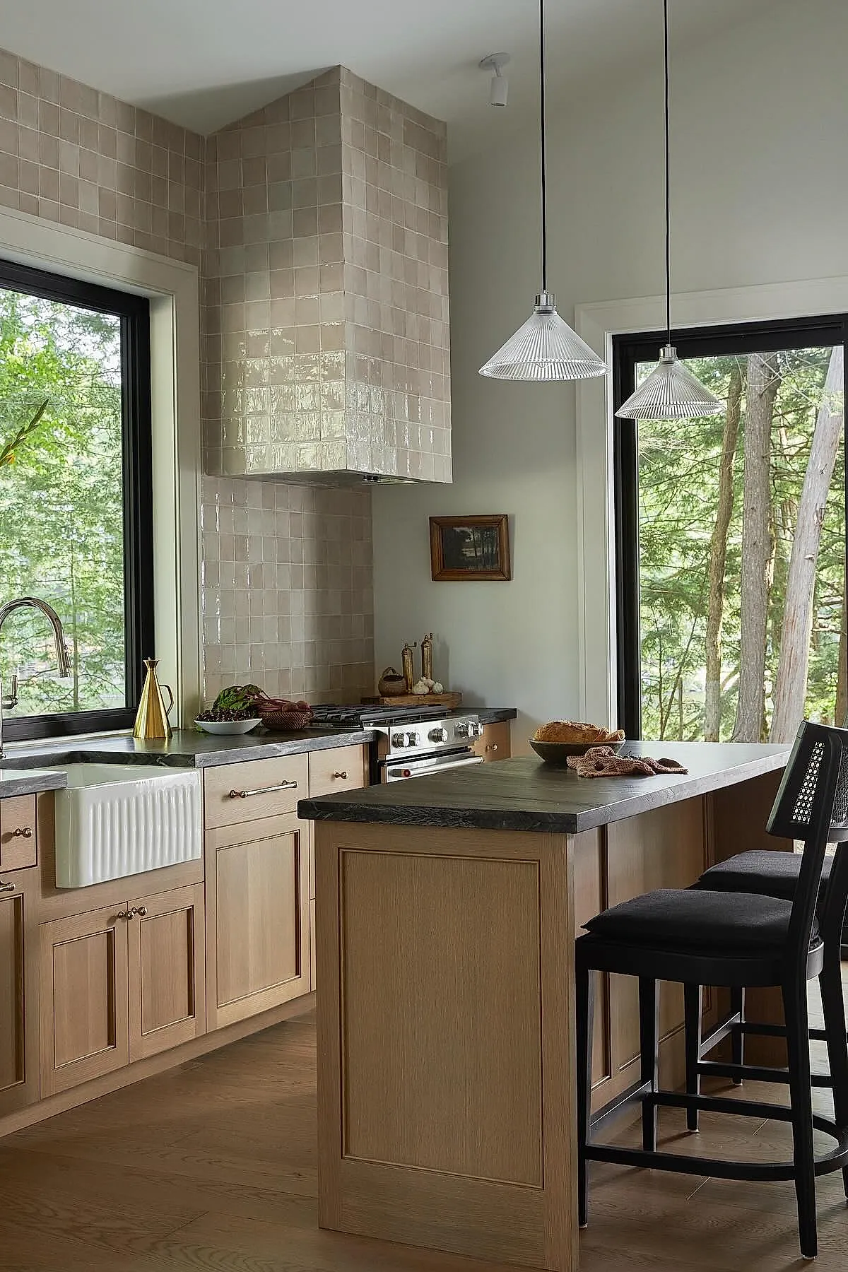 Cottage kitchen featuring glossy beige square tiles covering the backsplash and range hood, light wood cabinetry with brass handles, dark gray stone countertops, a white farmhouse sink, black framed windows showing green trees outside, and pendant lights hanging over a kitchen island with black stools.