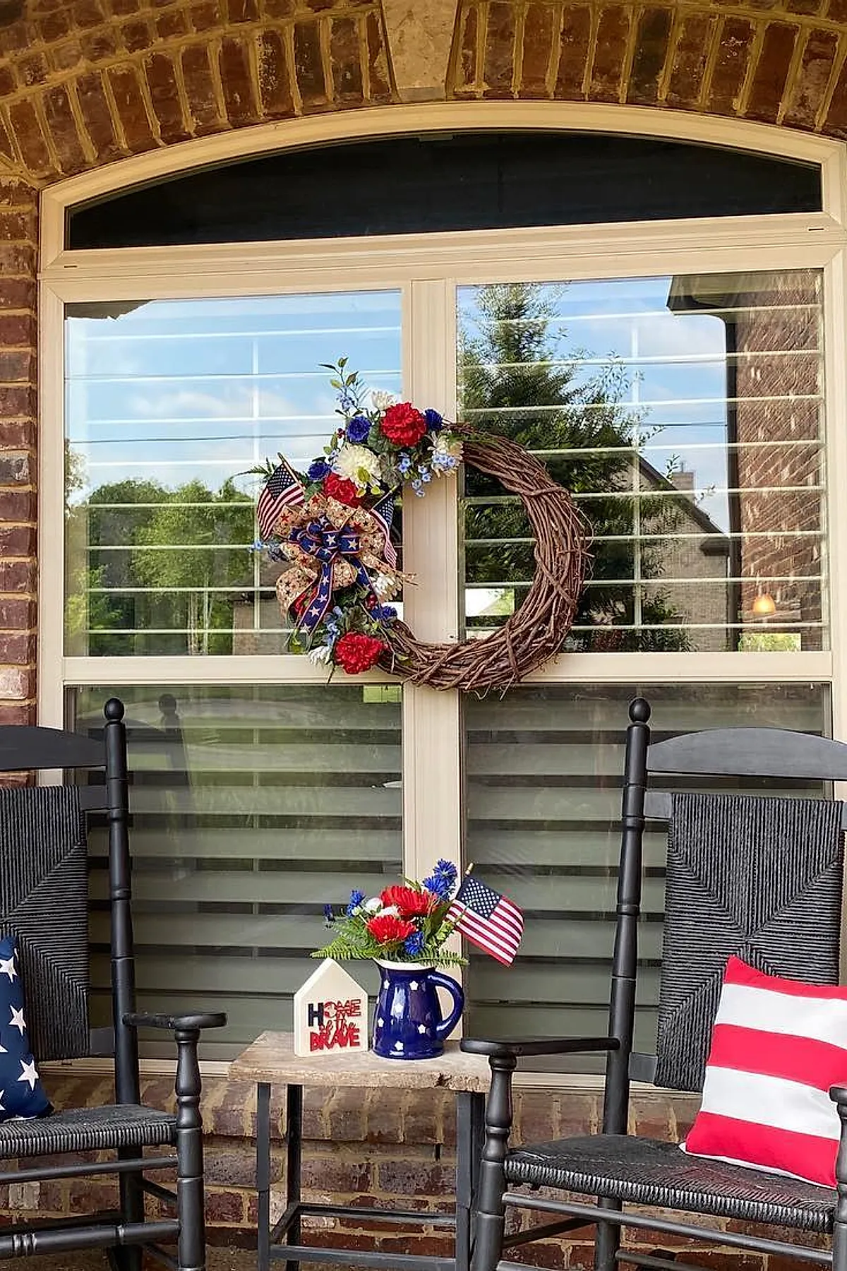 Patriotic wreath made of grapevine with red white and blue flowers and small American flags hanging on glass door surrounded by rocking chairs with patriotic cushions