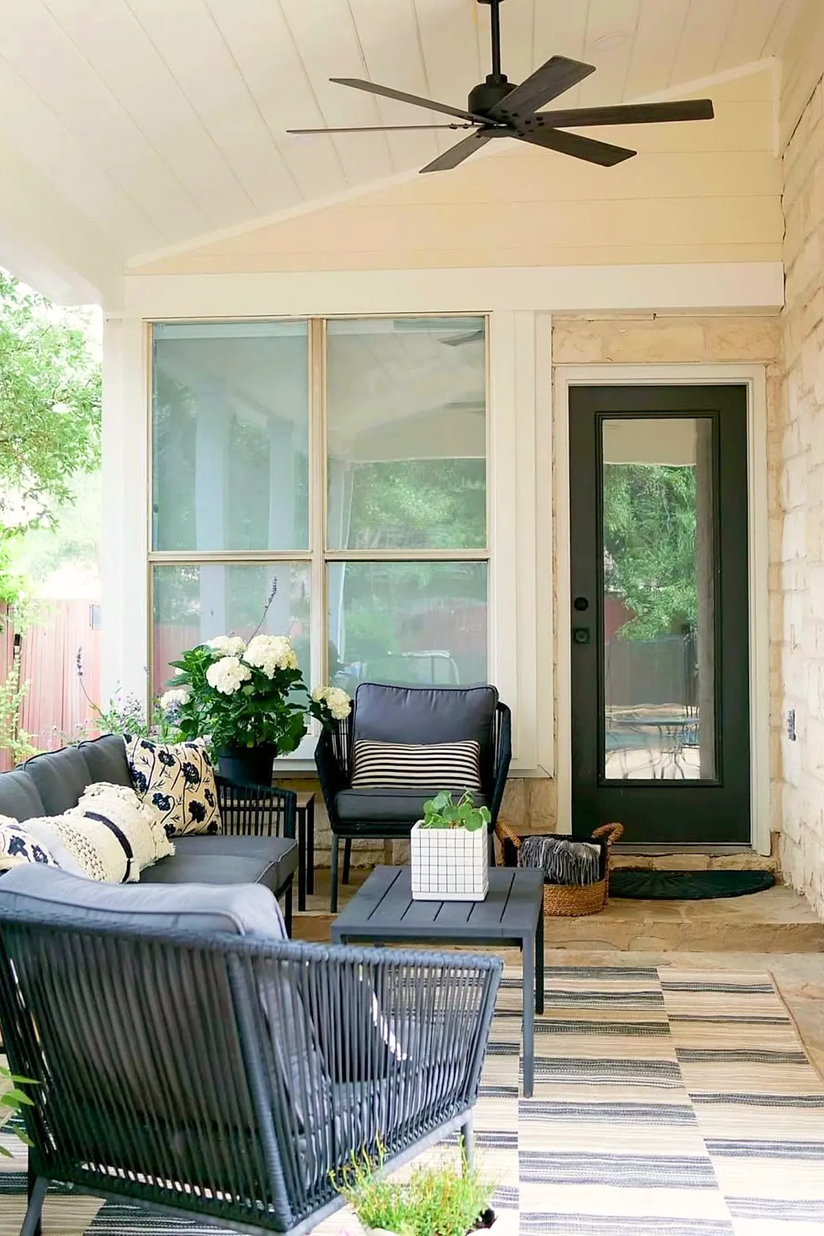 Covered patio attached to house featuring modern black metal framed seating with dark cushions and striped pillows, small black coffee table with white planter, potted hydrangea flowers, stone wall, ceiling fan, and large window overlooking green backyard.