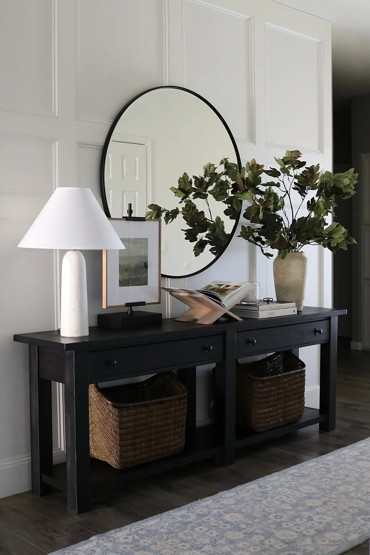 Modern entryway with a black wooden console table featuring drawers and woven baskets underneath, topped with a white lamp, framed artwork, an open book on a stand, and a large vase with green leafy branches; round black-framed wall mirror above the console; paneled white walls and dark hardwood flooring.