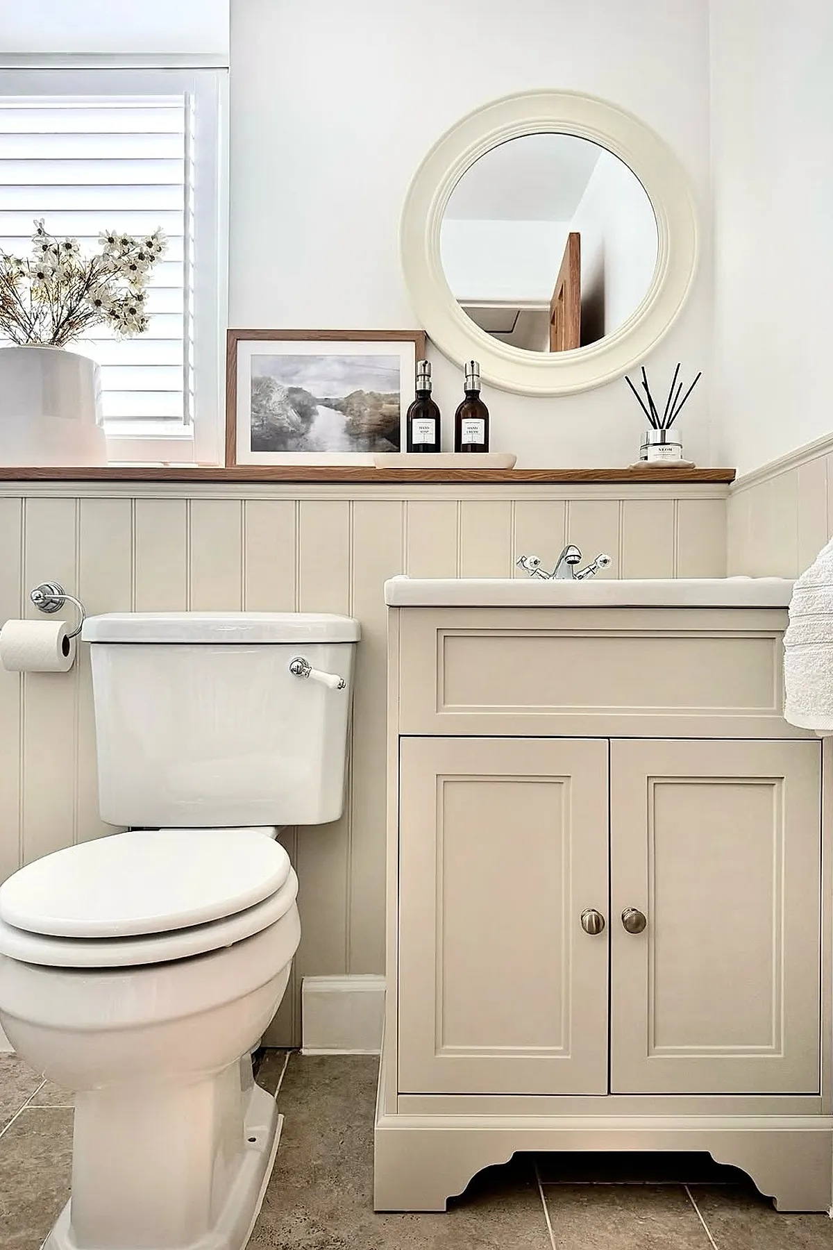 Warm neutral bathroom with cream colored vanity featuring paneled doors, a round cream-framed mirror above the sink, a white toilet, beige beadboard wainscoting on walls, and decorative items like a vase with white flowers and framed artwork on a wooden shelf behind the sink.