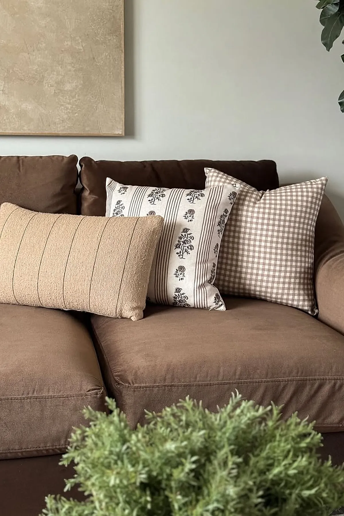 Close-up of a soft brown fabric sofa adorned with three decorative pillows: one beige textured lumbar pillow, one white pillow with black floral stripes, and one beige gingham check pillow, set against a pale wall with a neutral abstract painting.