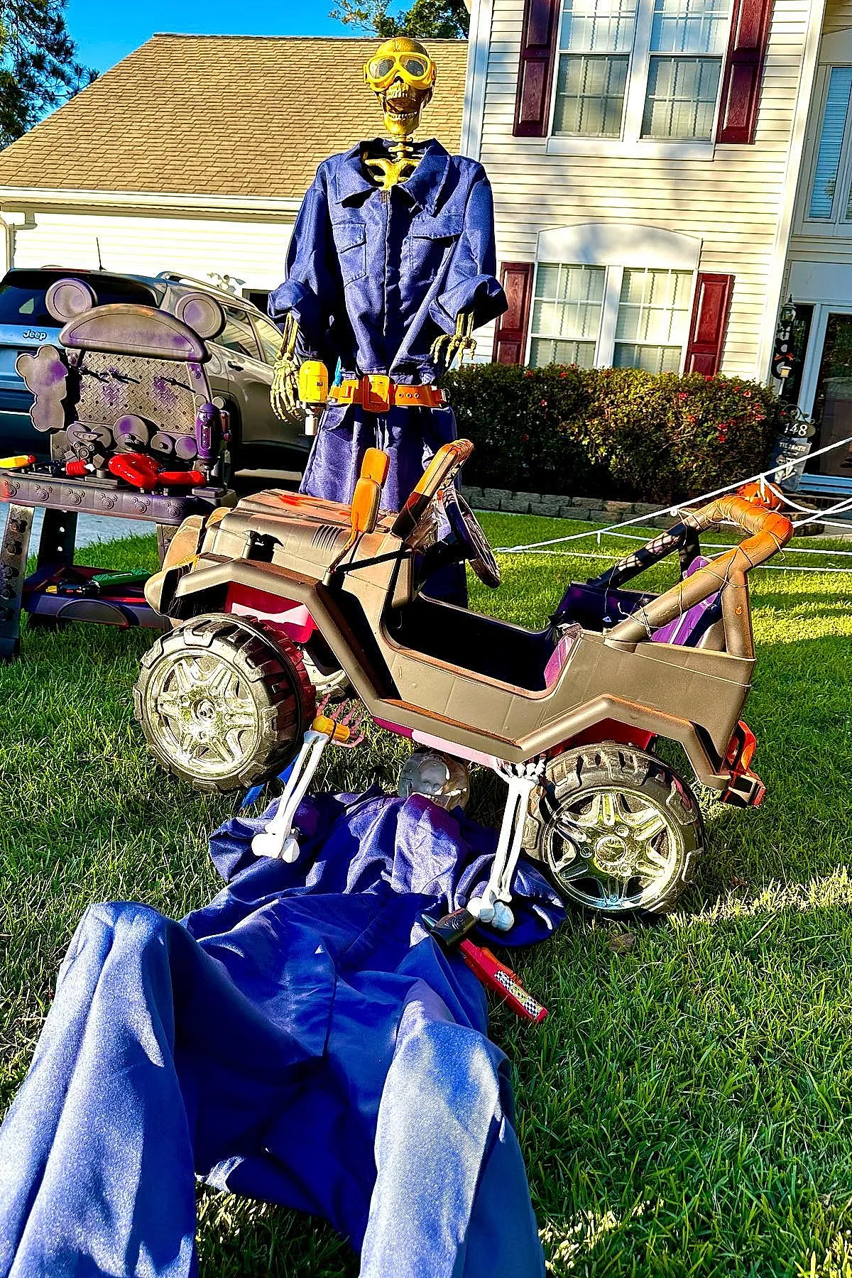 Skeleton figure wearing blue jumpsuit and yellow goggles standing behind toy jeep with another skeleton lying underneath pretending to fix it on green lawn in front of house.