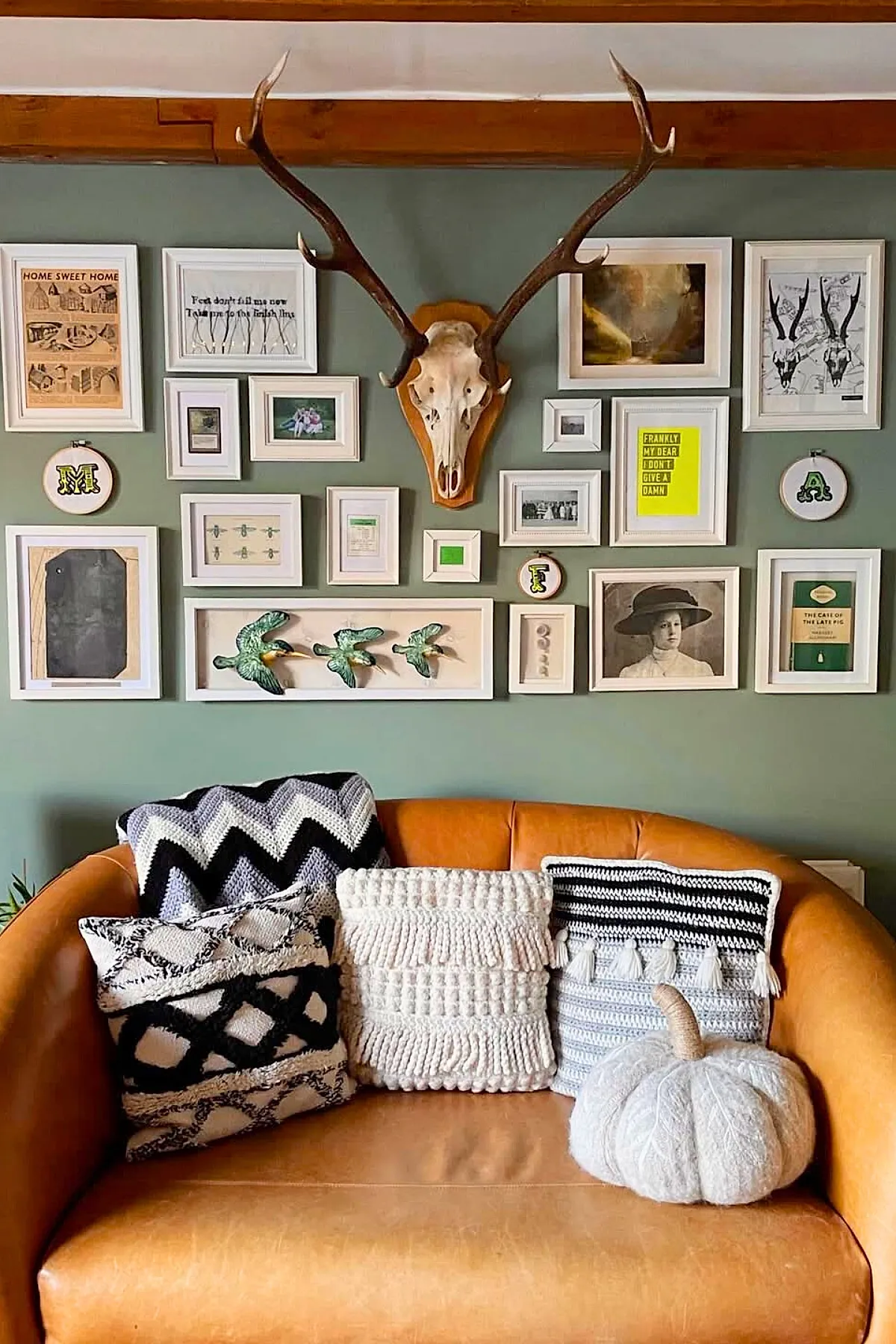 Living room wall decor featuring a mounted deer skull with large antlers surrounded by an eclectic gallery wall of framed artwork and embroidery hoops above a caramel leather sofa with textured black and white pillows and a knitted pumpkin cushion.