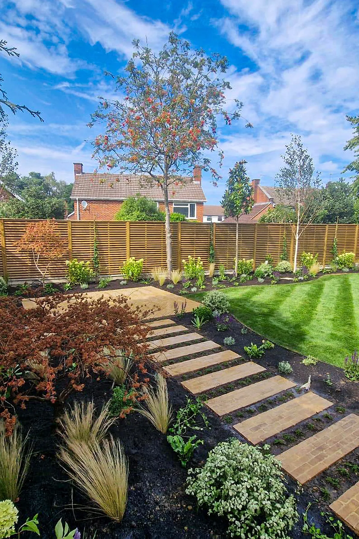 Landscape along fence line featuring a modern wooden slatted fence with a stepping stone pathway bordered by ornamental grasses, shrubs, and small trees in a garden setting