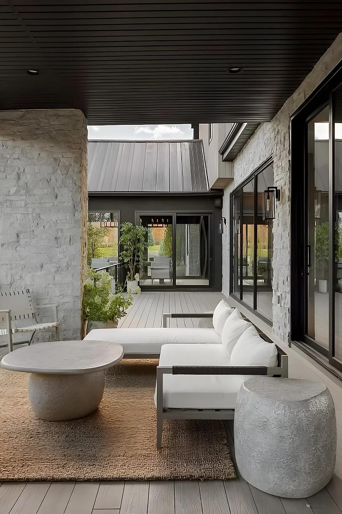Covered back patio attached to house with white cushioned modern sofa, stone side and coffee tables, dark paneled ceiling, light wood floor, and large windows.