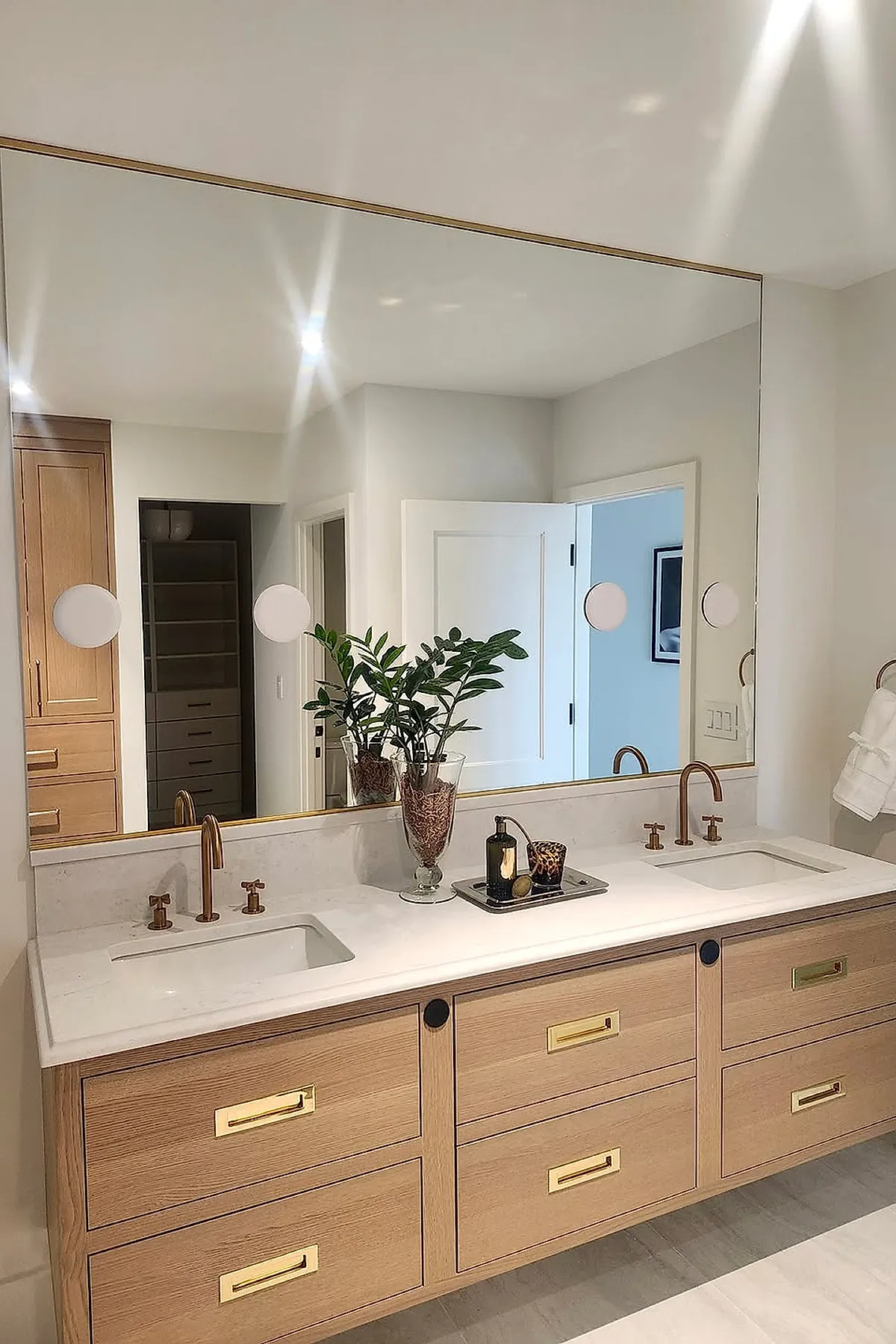 Large rectangular bathroom mirror with a slim brass frame above a white marble countertop featuring dual sinks and wood cabinetry with gold hardware, paired with brushed gold faucets and neutral walls.