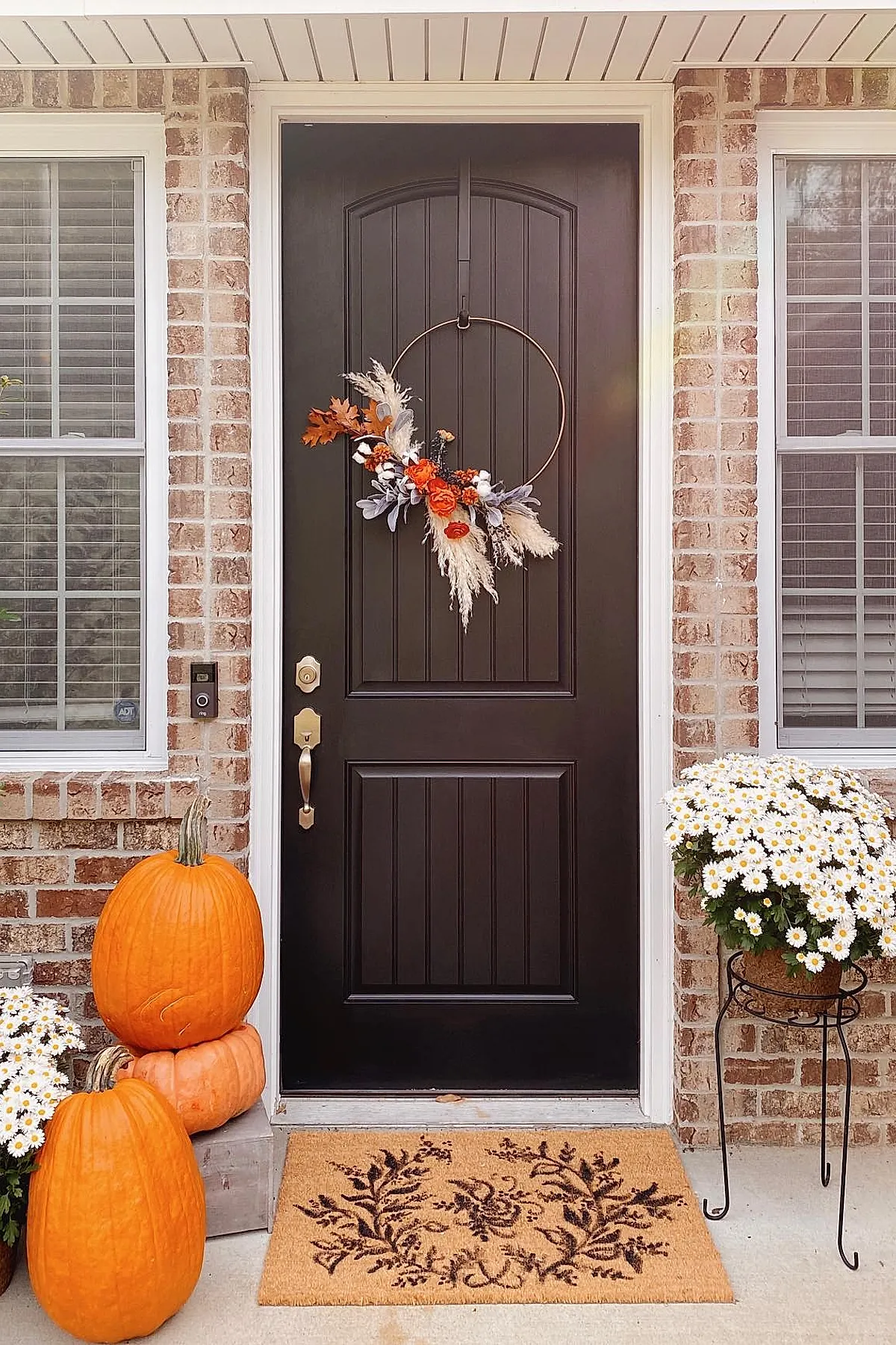 black front door with vertical panels featuring a minimalist fall wreath made from a bronze metal hoop decorated asymmetrically with dried pampas grass, orange flowers, gray leaves, and brown autumn leaves