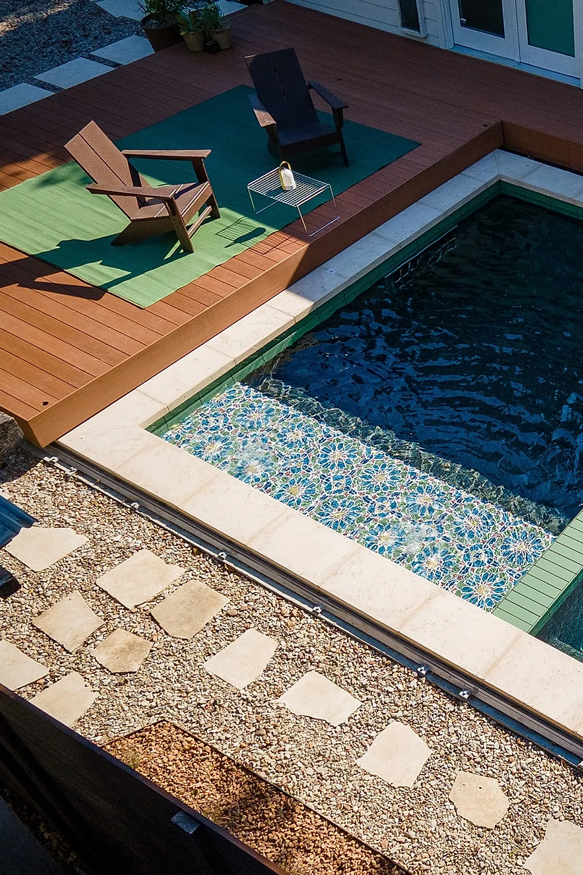 Small rectangular plunge pool with intricate blue, green, and white mosaic tiles in the shallow area, bordered by cream-colored stone edging and a wooden deck featuring two dark brown Adirondack chairs on green outdoor rugs.
