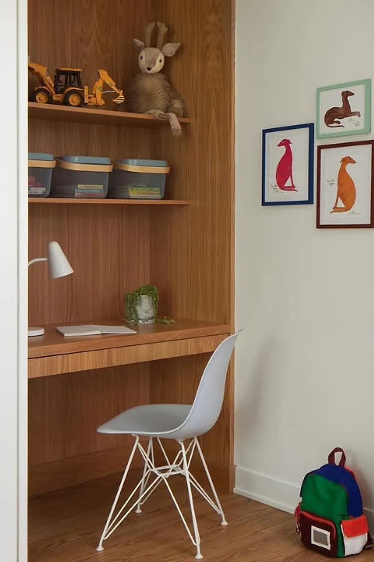 Kids desk area in bedroom featuring a built-in wood kids study desk with matching shelves, a gray modern chair with white metal legs, plastic storage bins on upper shelf, a white desk lamp, a small green plant on the desk, and three framed animal prints on the adjacent light-colored wall.