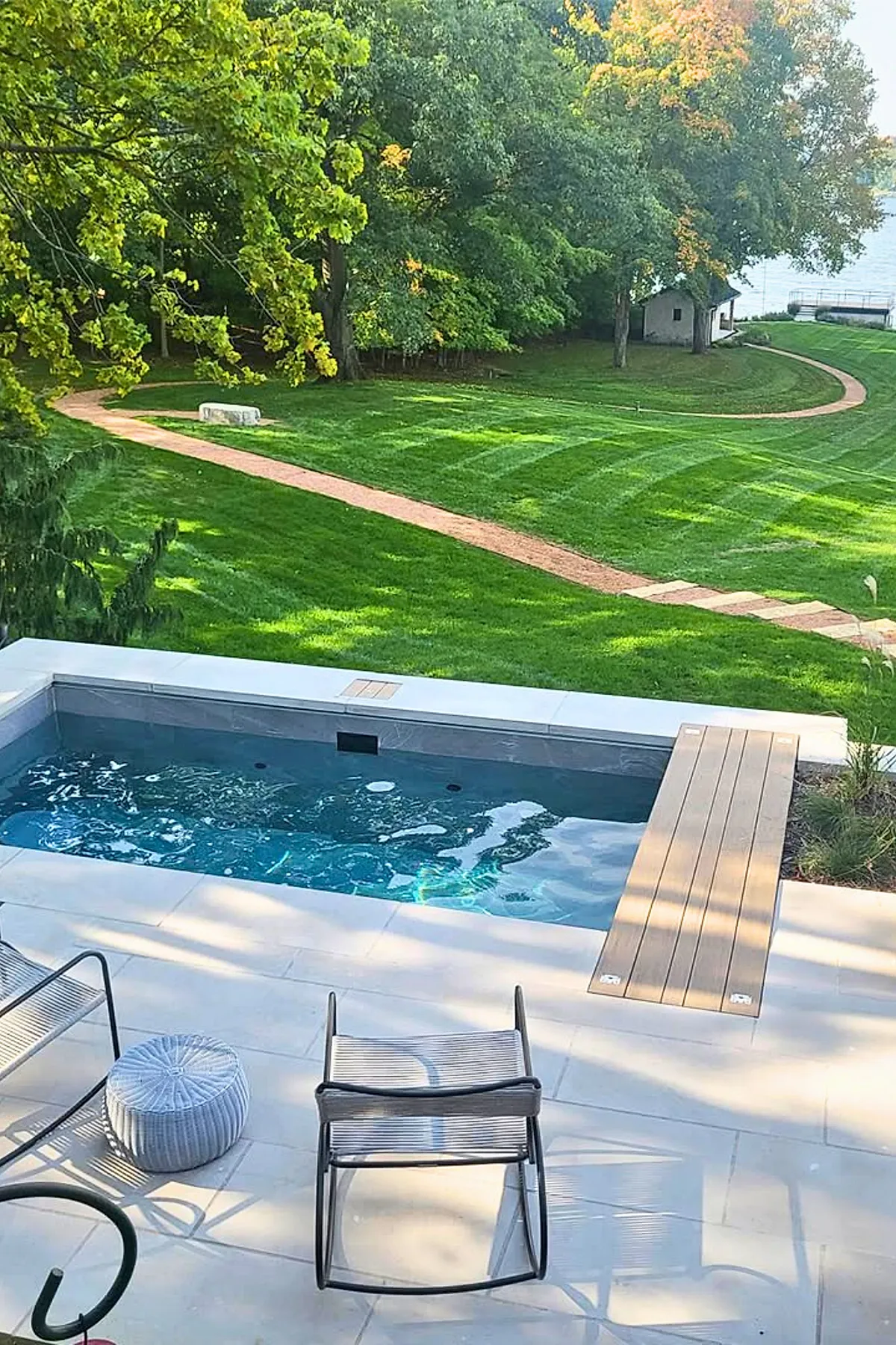 A small modern inground plunge pool with bubbling water, surrounded by light stone tiles and two metal lounge chairs, overlooking a lush green lawn with winding path and tall trees in the background.