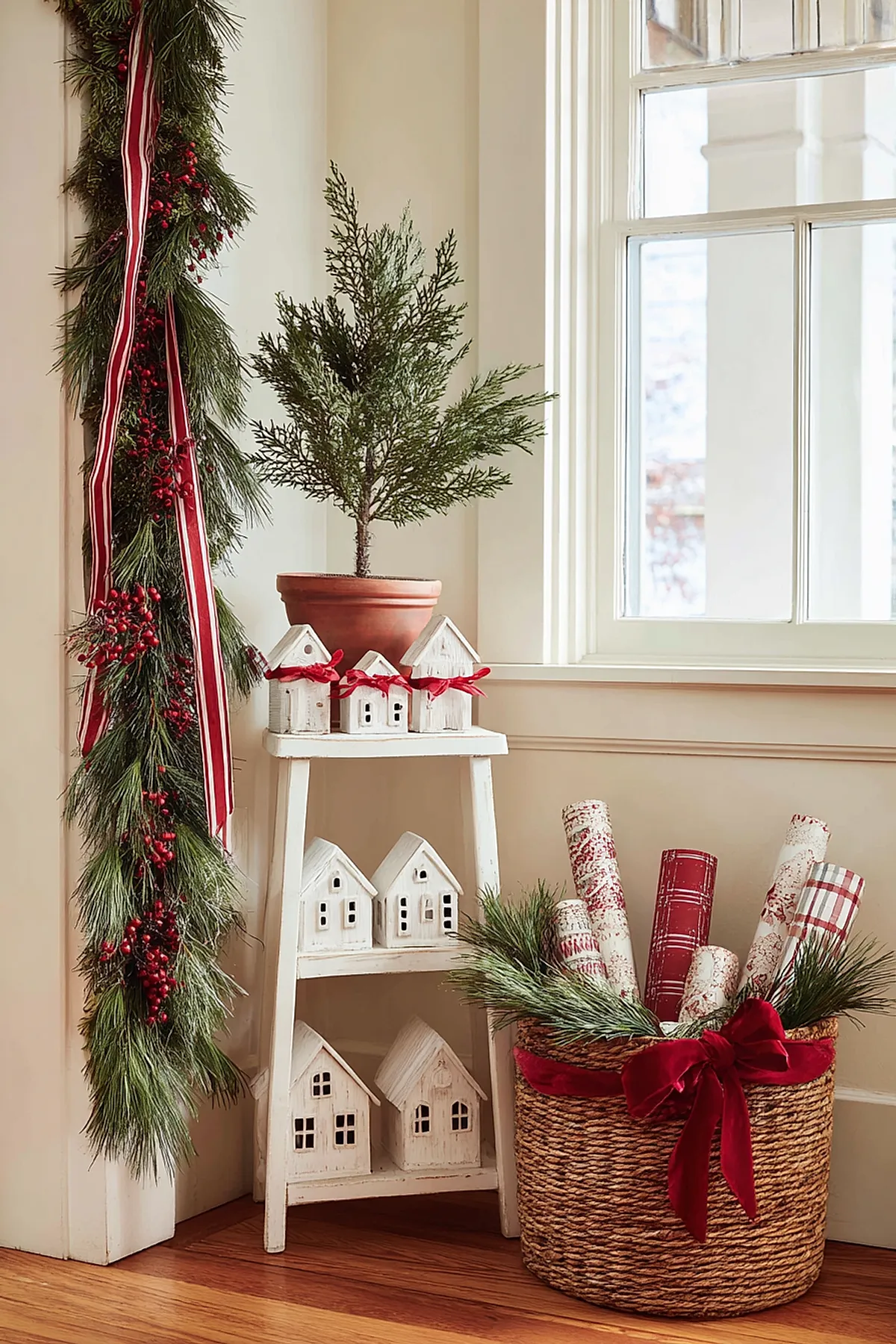 Corner vignette featuring a mini evergreen tree surrounded by white ceramic houses and a festive basket filled with wrapping paper and velvet bows for vintage inspired christmas decor.