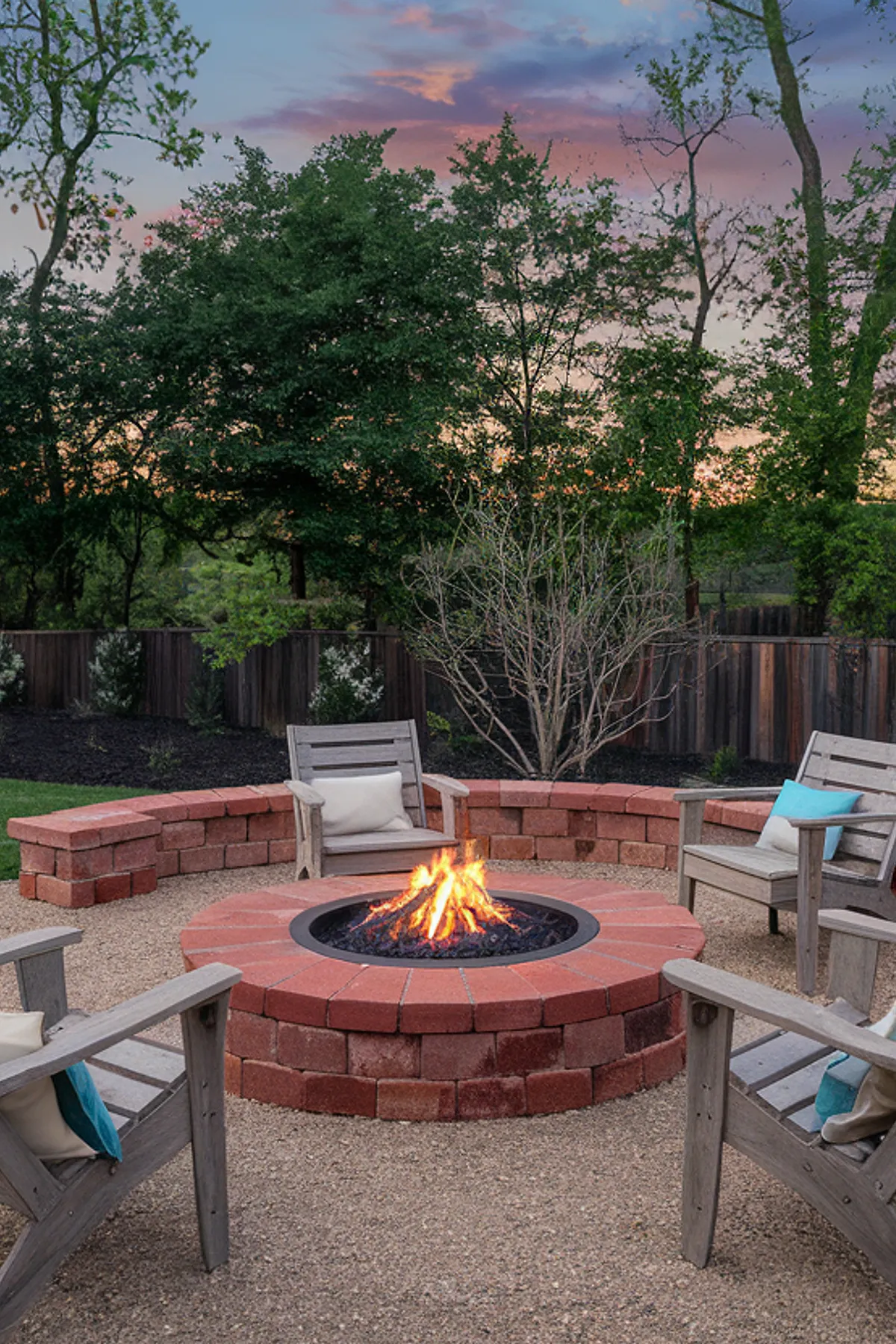 A rustic brick fire pit surrounded by wooden chairs in a backyard setting.