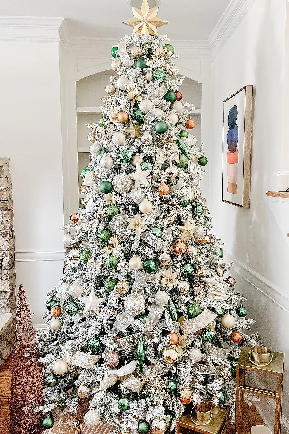 White flocked Christmas tree densely decorated with green, gold, rose gold, cream baubles, sheer ribbon, star ornaments, and a large golden star topper in a bright room next to metal wire trees and brass lanterns.
