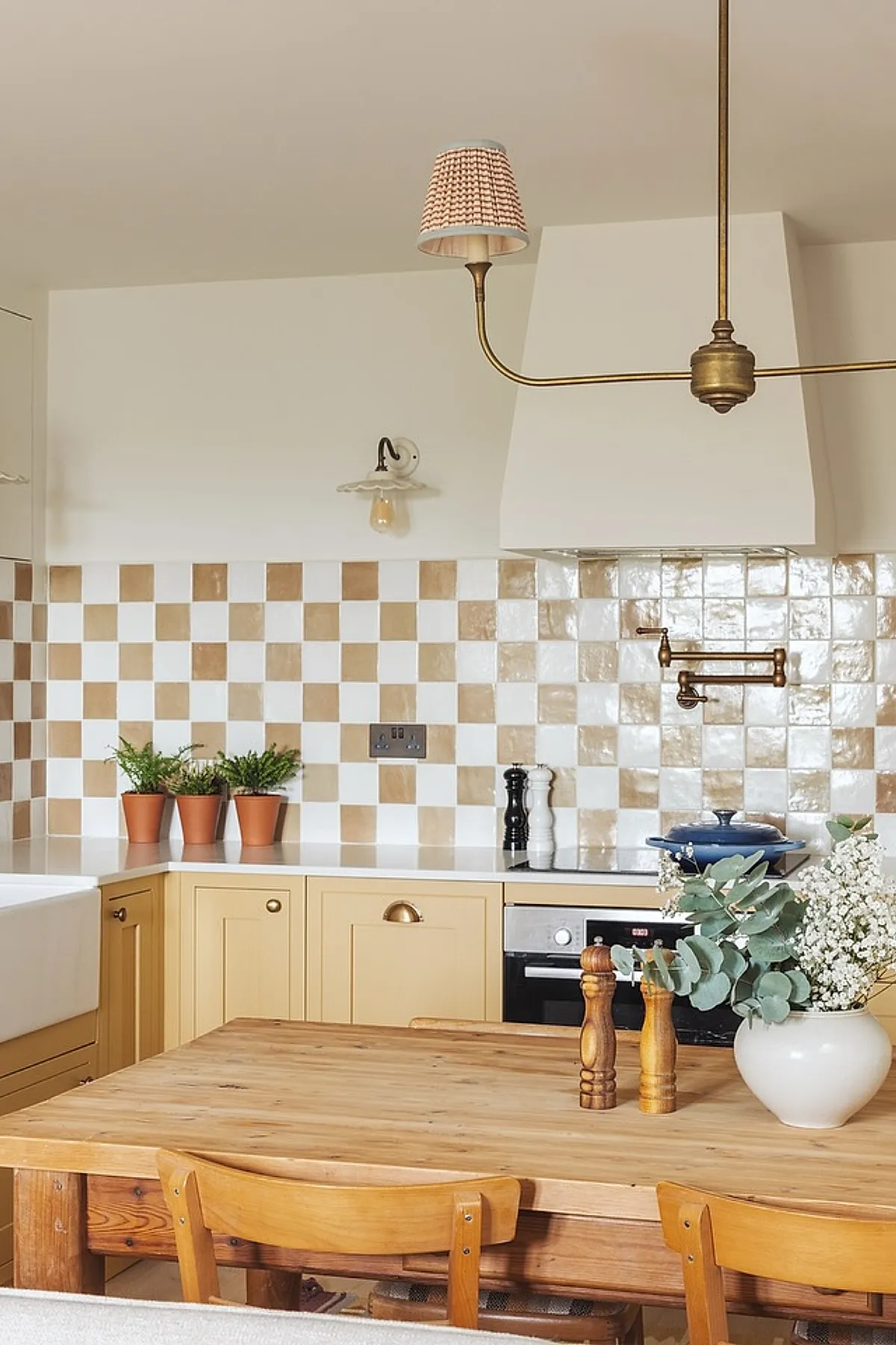 Yellow cottage kitchen featuring light yellow cabinetry with brass hardware, a white and beige checkerboard tile backsplash, white walls above, built-in oven, wooden dining table with natural finish, three small terracotta pots with green plants on the countertop, and brass wall sconces.