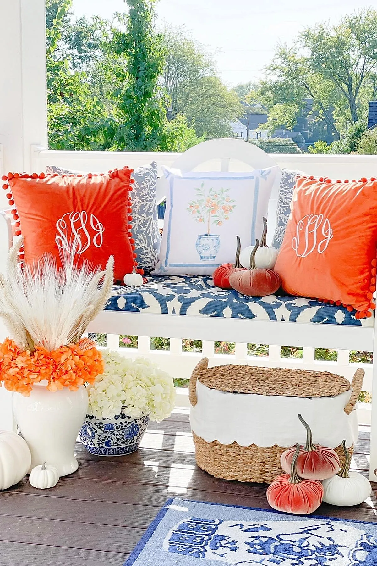 white wooden bench with blue and white patterned cushion, two bright orange pompom trim pillows, one floral pillow, small white and coral velvet pumpkins on seat, wicker basket with scalloped cloth liner, white ceramic vase with dried pampas grass and orange flowers, blue and white planter with white flowers