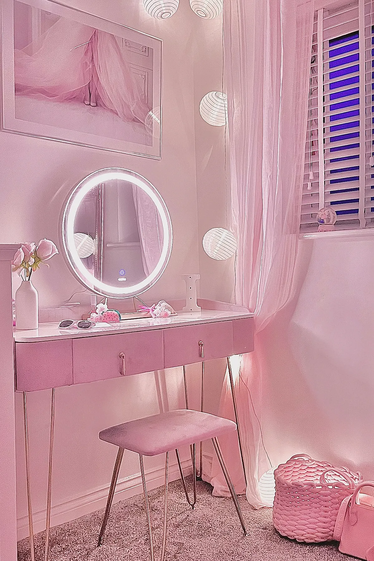 pink bedroom vanity with round LED-lit mirror on desk, soft pink cushioned stool with gold legs, white vase with pink flowers on surface, sheer pink curtains covering window with white blinds, illuminated paper lantern string lights hanging near wall