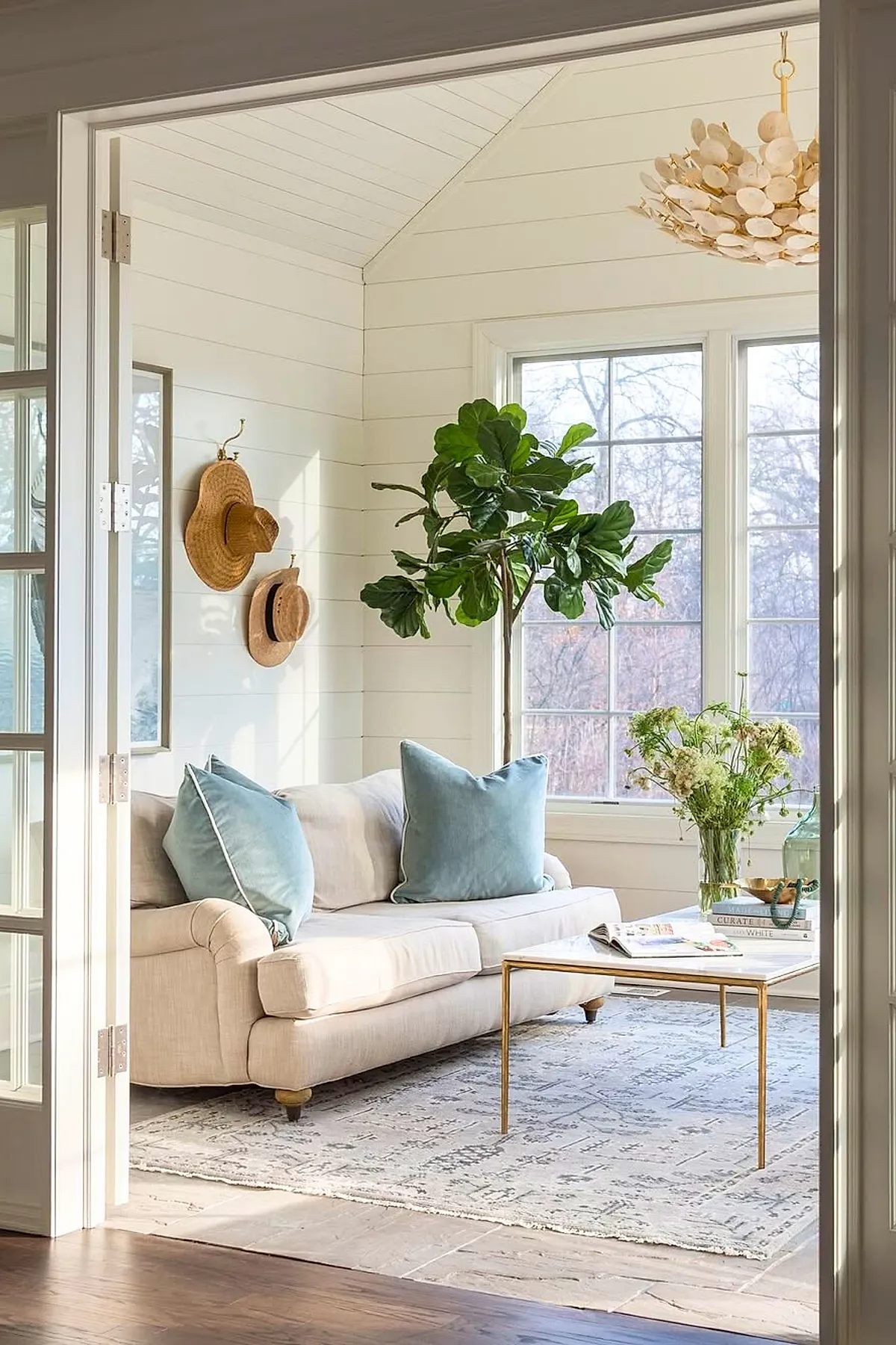 Sunroom with beige sofa accented by light blue pillows, a tall green plant by large windows, white shiplap walls, a light patterned rug, gold-legged white coffee table holding books and a glass vase with flowers, under a vaulted ceiling.