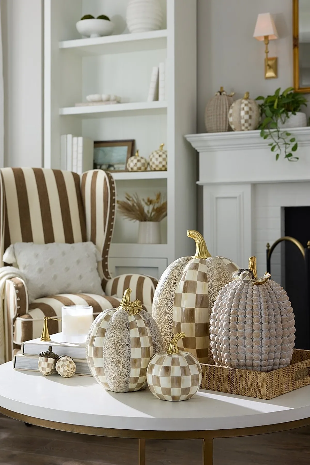 Round white coffee table displaying various sizes of beige and cream checkerboard patterned pumpkins with gold stems, alongside a textured pumpkin in similar tones, set in front of a striped armchair and white fireplace mantel decorated with matching pumpkins and greenery.