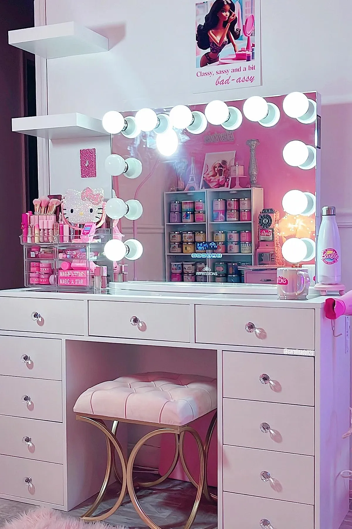 white vanity desk with multiple drawers, large mirror framed by round white light bulbs, tufted pink cushioned stool with gold metal legs, clear acrylic makeup organizers holding pink brushes and lipsticks, pink wall accents and Barbie-themed decor