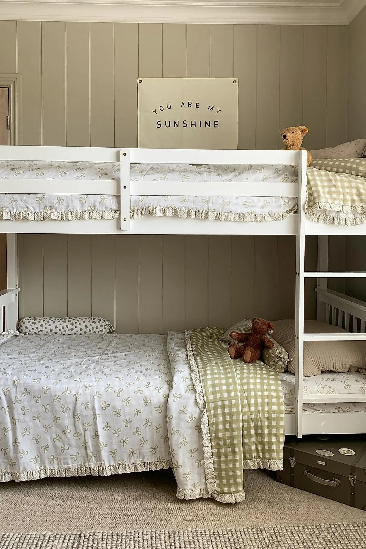 White bunk bed with beige wall paneling background. Bedding features light cream fabric with small beige bows and ruffled edges, paired with green gingham plaid blankets. Two teddy bears sit on the beds, one at each headboard end. Beige carpet underfoot and stacked dark suitcases near ladder.
