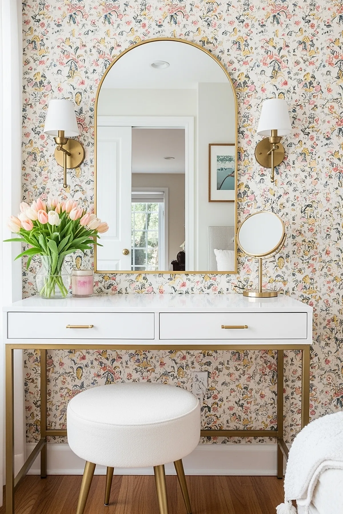 white vanity table with gold legs and handles, large arched gold-framed mirror mounted on floral wallpaper, two gold wall sconces with white shades on each side of mirror, clear glass vase with pale pink tulips, round white upholstered stool with gold legs