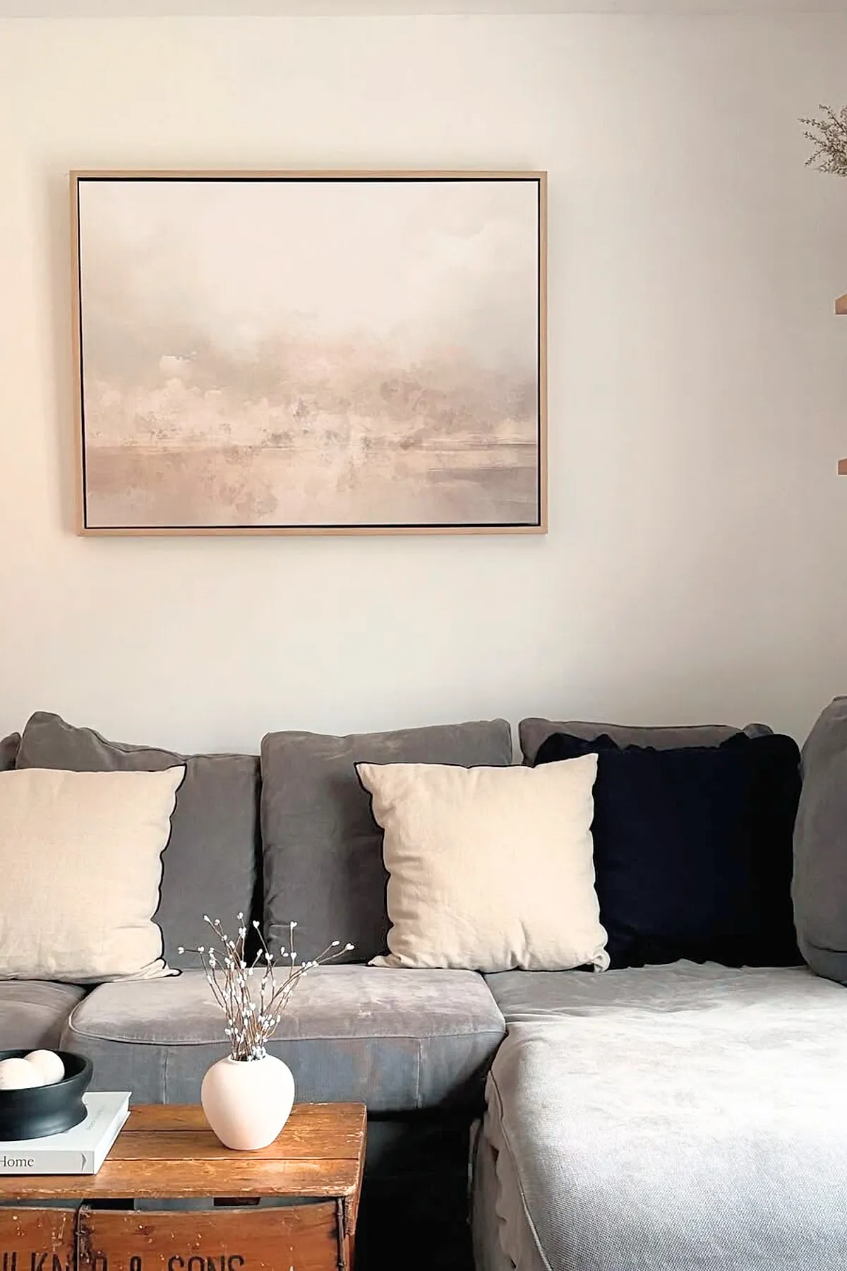 Earthy living room featuring a gray sectional sofa with neutral cream and black pillows, a rustic wooden coffee table topped with a small white vase holding delicate dried branches, and an abstract beige and blush wall art hanging above the sofa on a light-colored wall.