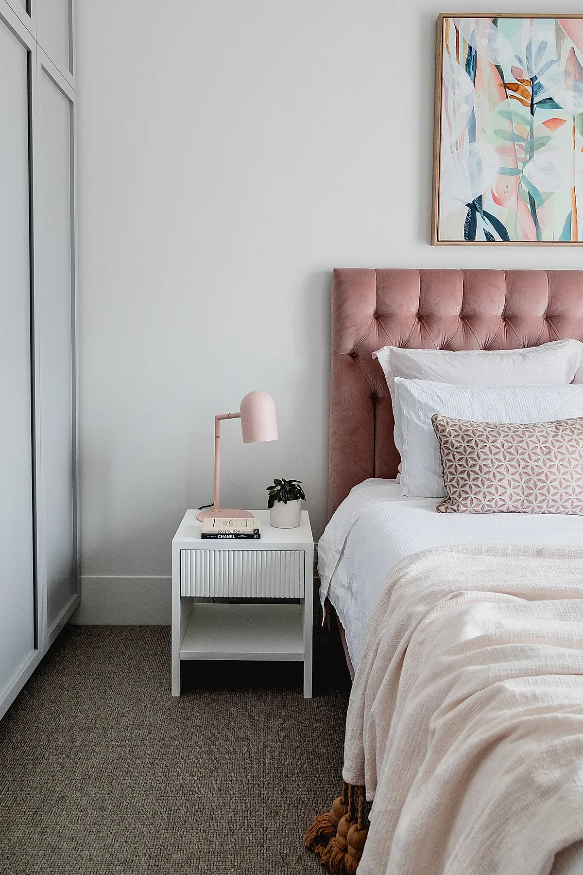 teen girl bedroom featuring a dusty pink velvet tufted headboard, white bedding with textured blush throw blanket, geometric patterned pillow, white minimalist nightstand with a pink desk lamp, small potted plant, and abstract pastel wall art above the bed.