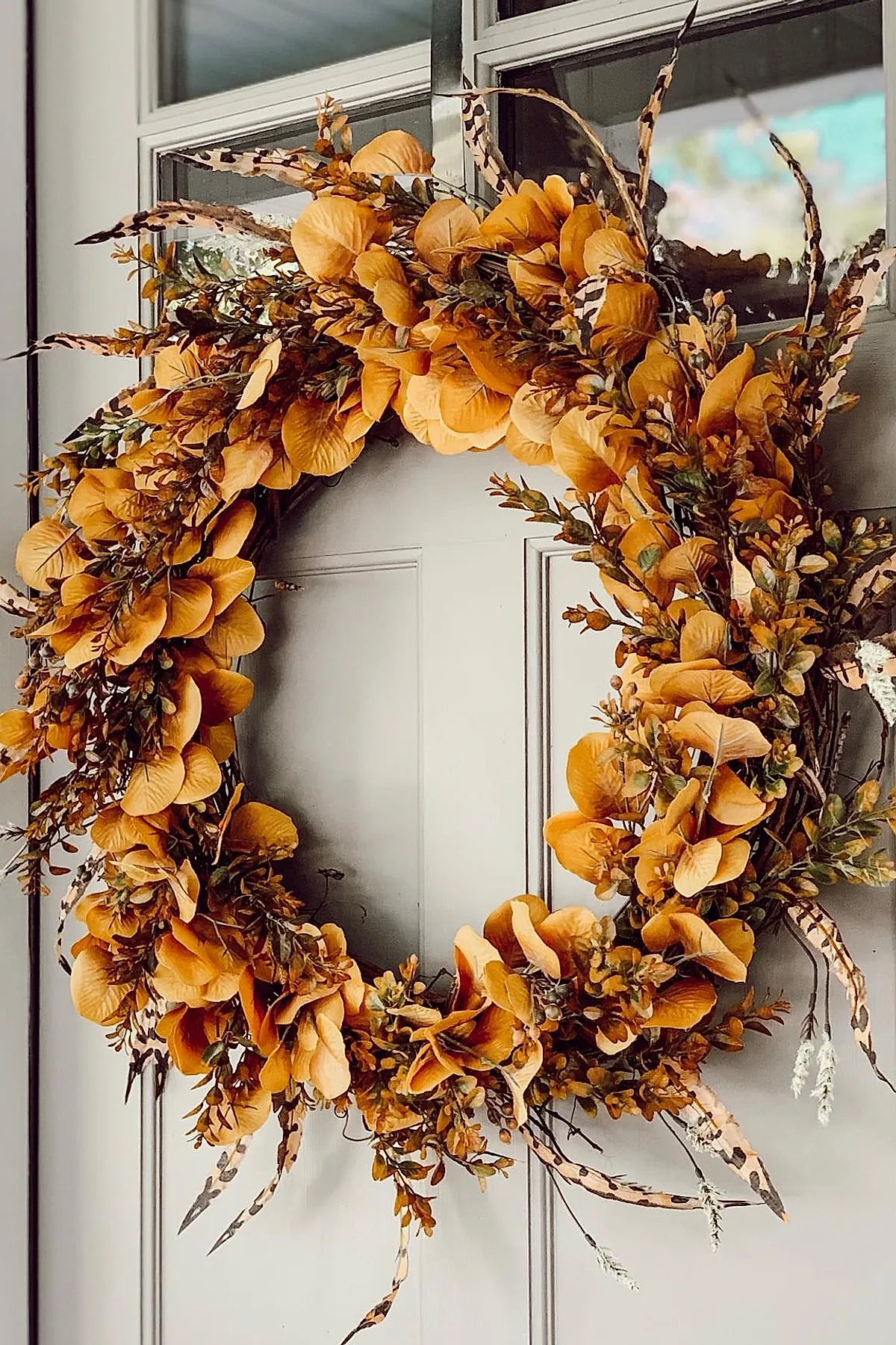fall wreath featuring mustard yellow leaves mixed with green sprigs and long brown spotted feathers arranged on a gray paneled door