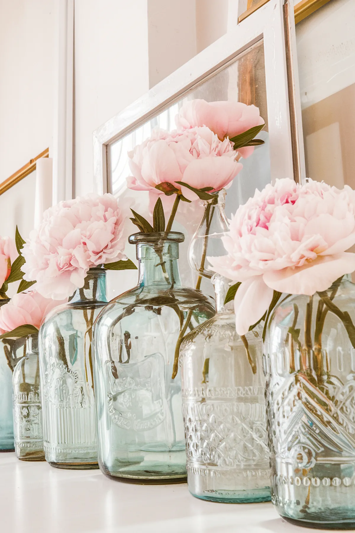 A mantel decorated with vintage glass bottles holding pink peonies.