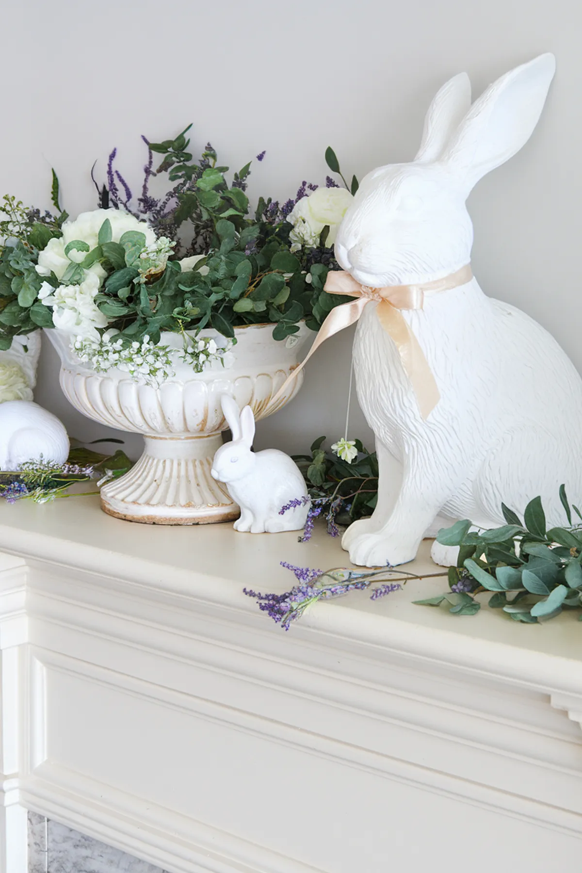A spring mantel decorated with white rabbits, greenery, and floral arrangements in an urn.