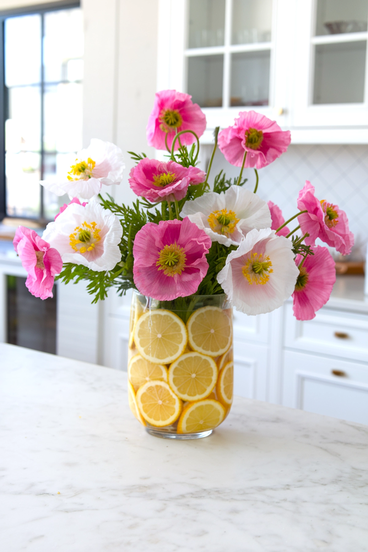 A vibrant summer floral arrangement featuring pink and white poppies with yellow centers, arranged in a tall clear glass vase filled with fresh lemon slices. The bouquet sits on a white marble countertop in a bright kitchen with white cabinets and gold hardware, creating a cheerful and refreshing atmosphere.