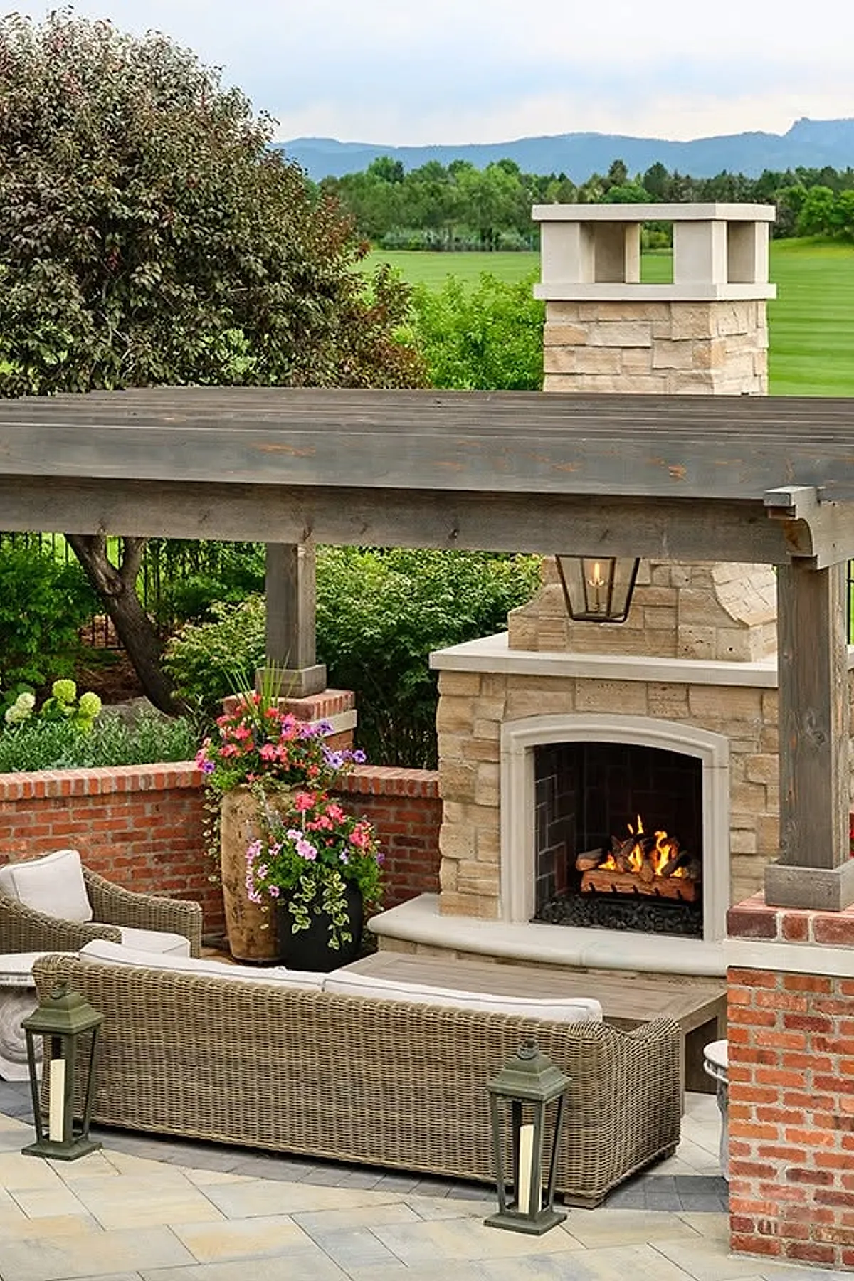 Outdoor covered patio with large stone chimney fireplace featuring beige stonework and a wooden mantel, surrounded by wicker sofas with white cushions, brick half walls, colorful flowering plants in pots, and greenery beyond the patio.